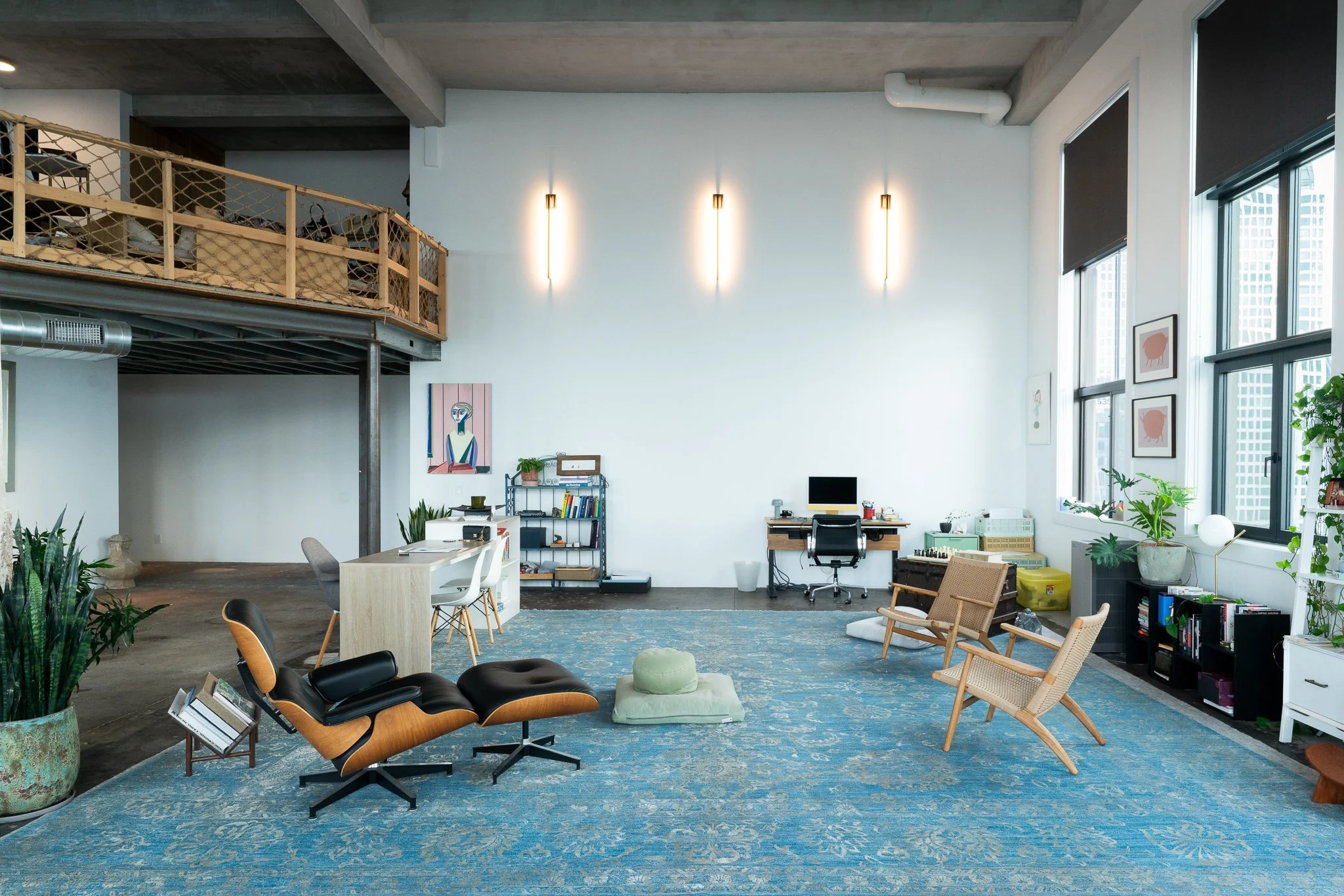 A spacious, modern interior with high ceilings, large windows, and a mix of seating options. There's a blue patterned rug on the wooden floor, plants, artwork on the walls, and a workspace with a desk and chair. The space has an industrial touch with visible ductwork and a second-floor balcony with wooden railings.