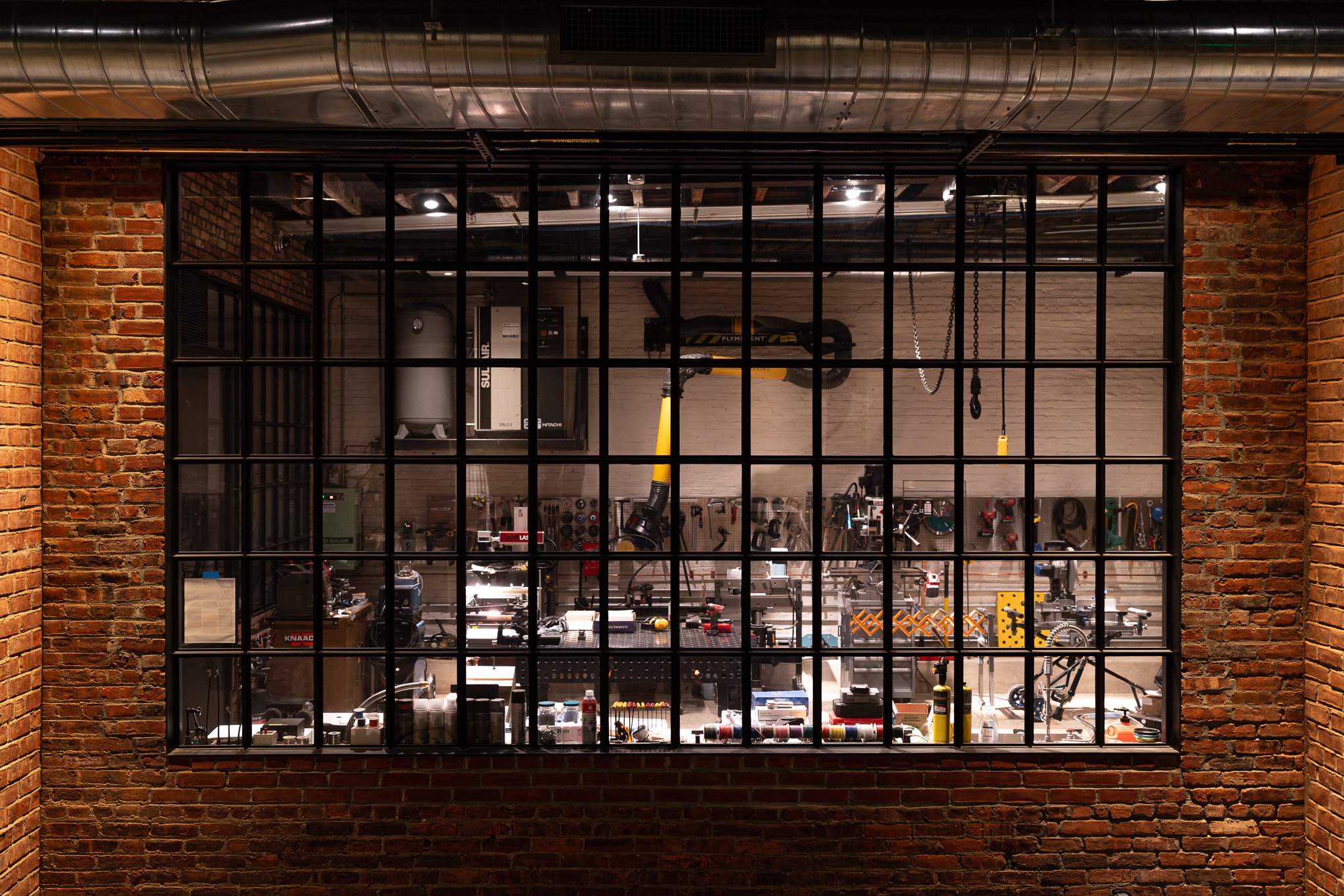 View through a large industrial-style window into a workshop with tools, equipment, and supplies, surrounded by brick walls and exposed ductwork on the ceiling.