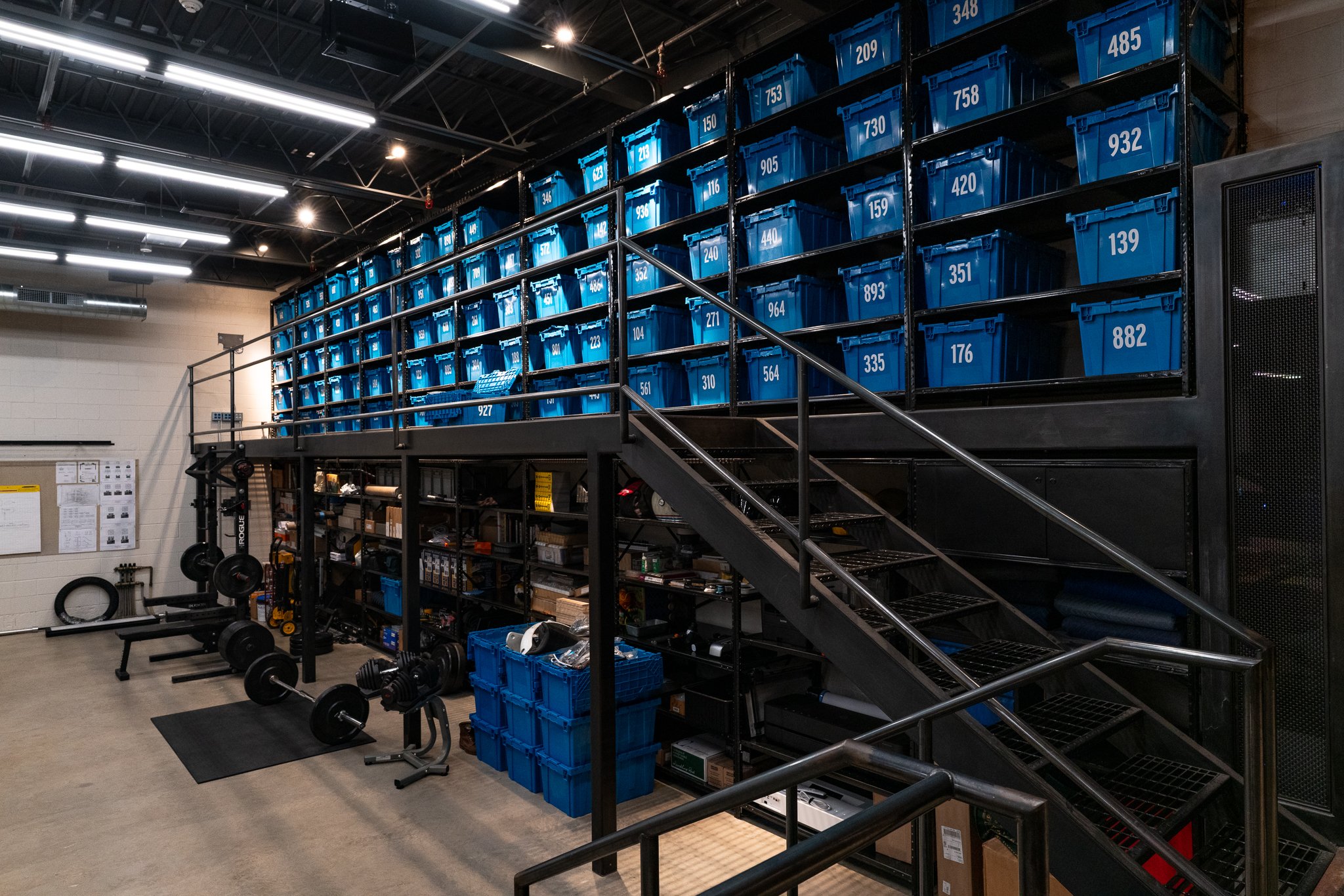 Gym with black weightlifting equipment, blue storage bins on shelves, and stairs leading to a second level with more bins. Gym lighting and ceiling visible.