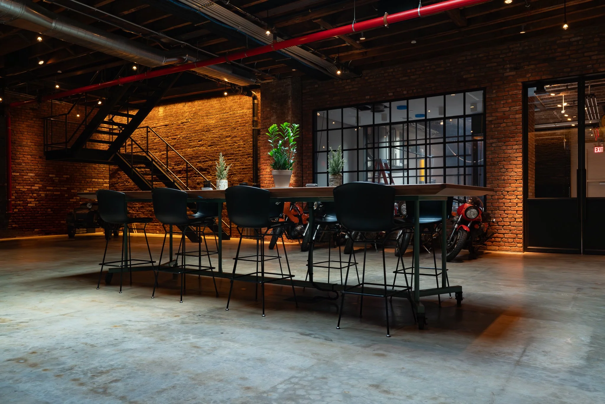 Interior of a modern industrial-style office or coworking space with brick walls, a staircase, tables, chairs, and potted plants.