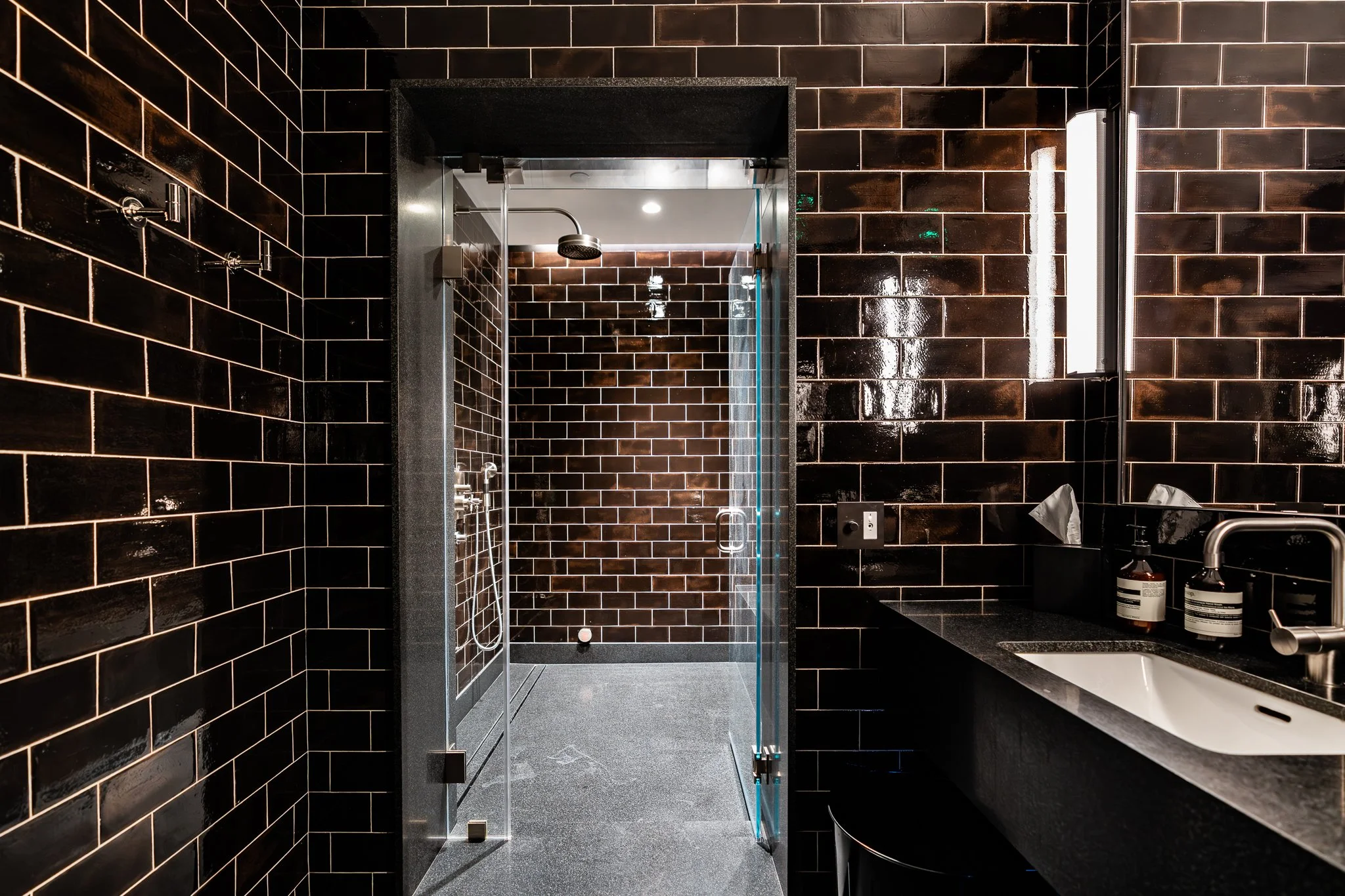 A modern bathroom with black tiled walls, a glass shower door, a rainfall showerhead, a black countertop with soap bottles, and a white sink.