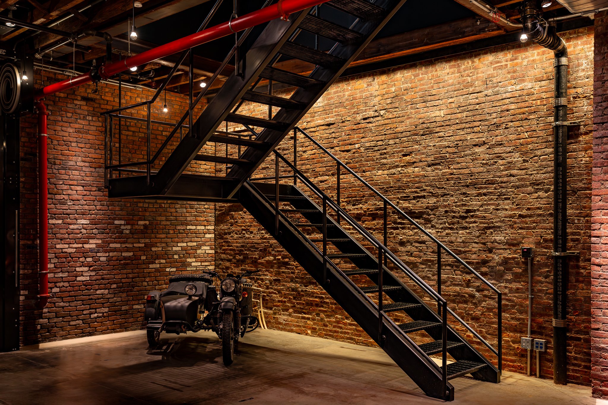 A black metal staircase with a railing against a brick wall interior. Below the stairs, a small motorcycle is parked in a room with a dark wood floor and exposed pipes on the ceiling.