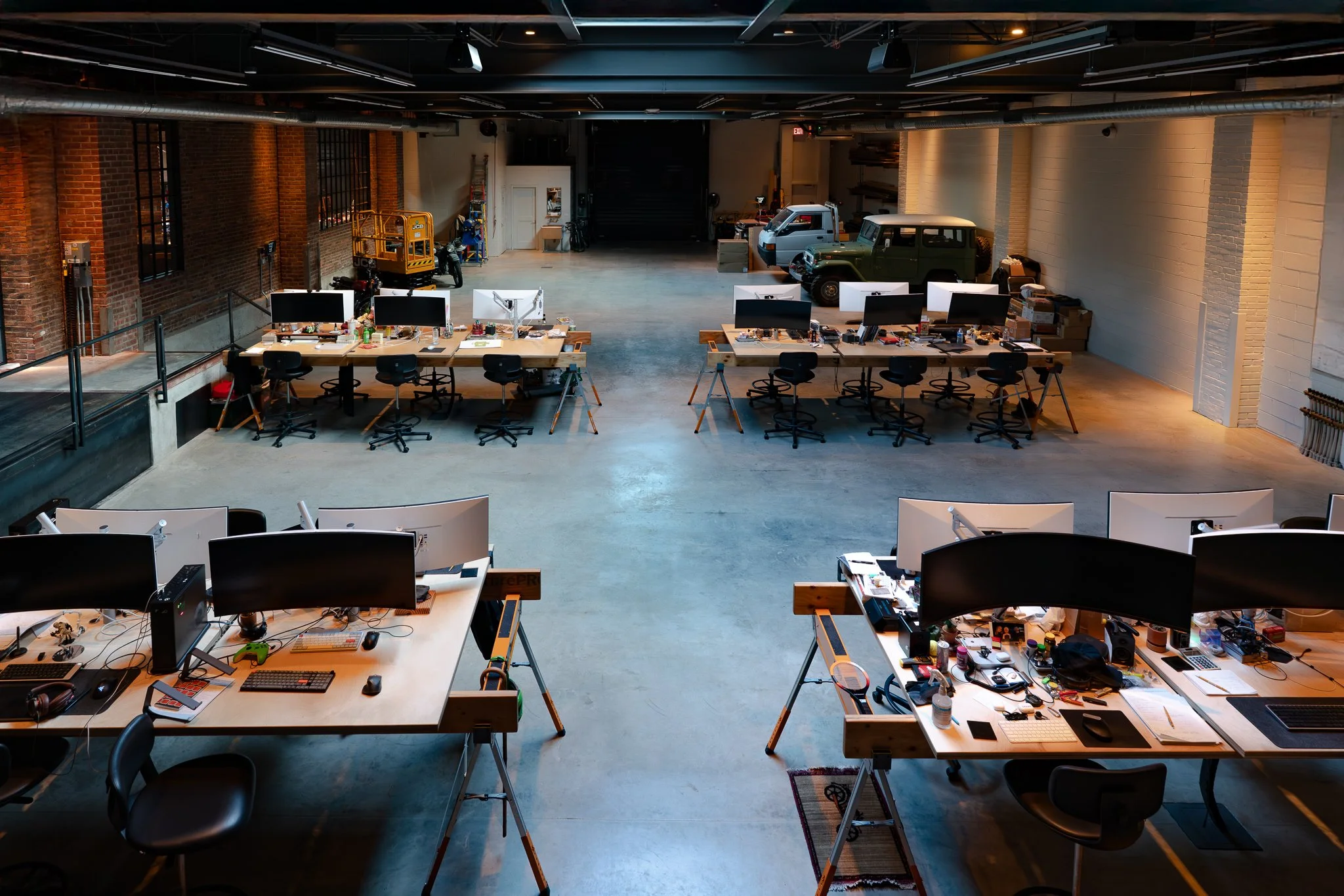 An industrial-style office space with wooden desks, computer monitors, and chairs. The space includes a brick wall, large windows, and vehicles like a golf cart and two trucks at the back. The area appears to be used for creative or technical work.