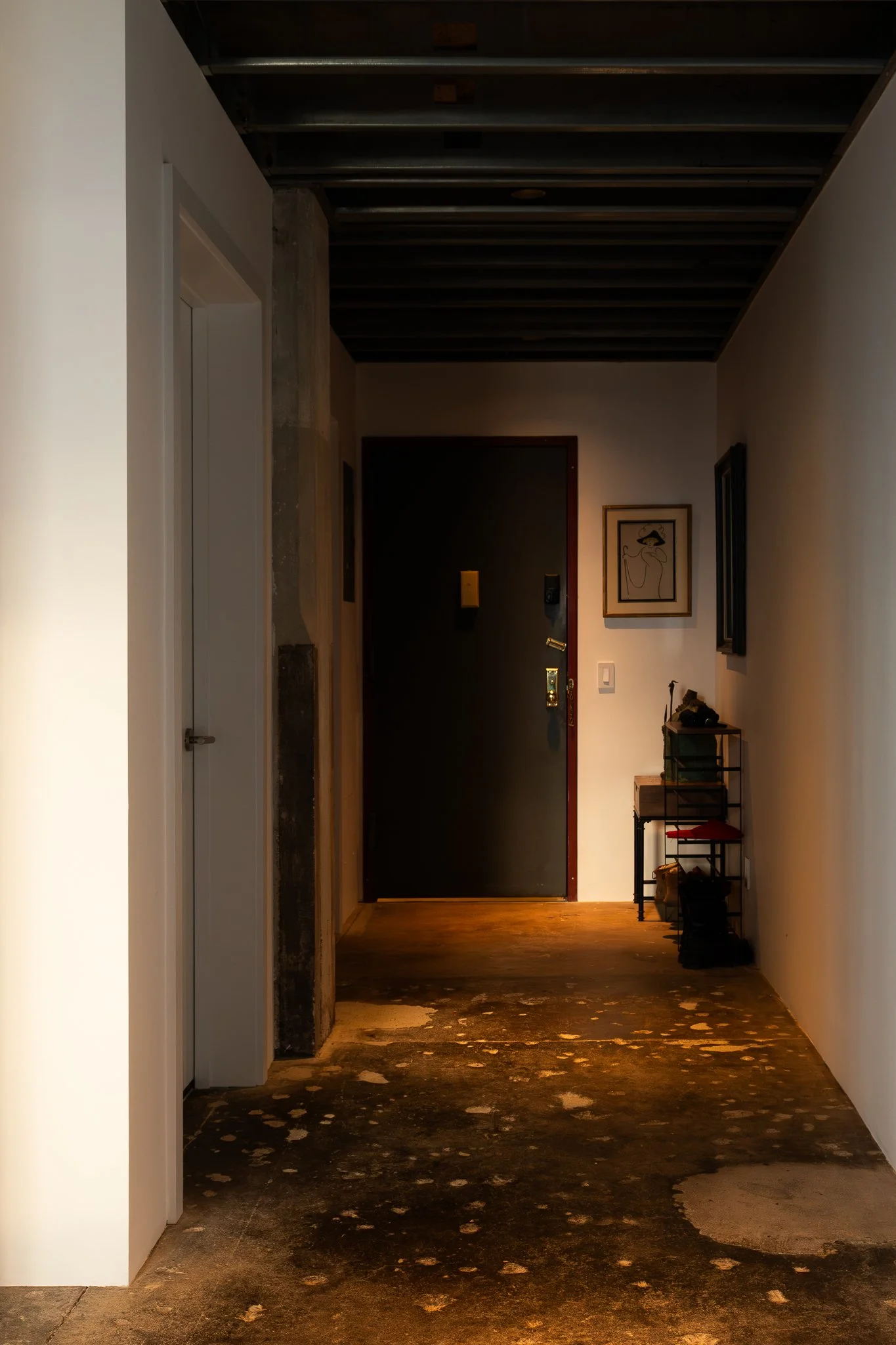 A narrow hallway with a dark front door, framed artwork on the walls, a small table with decorative items, and a worn wooden floor with scattered paint splatters.