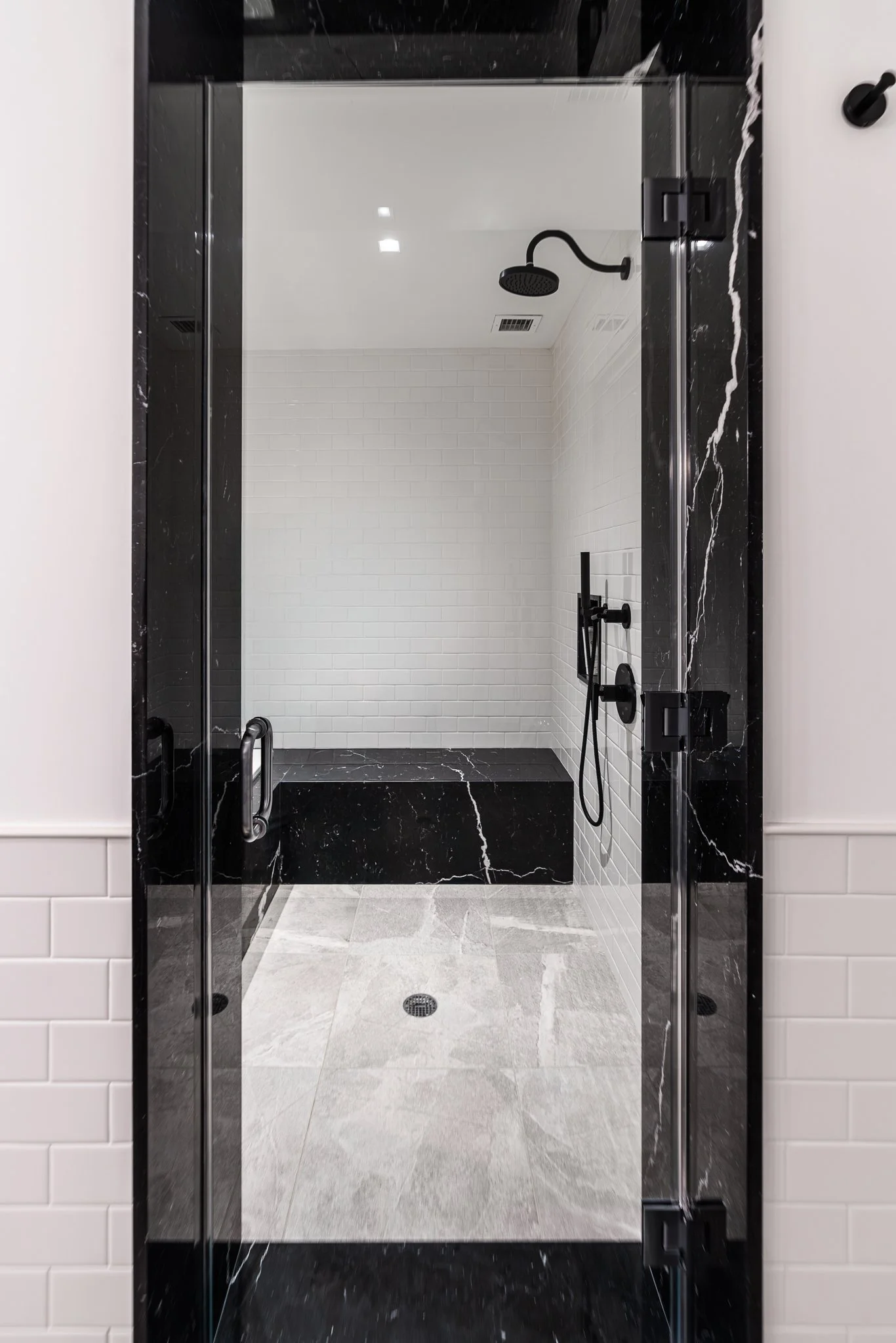 A glass shower door with black marble trim, showing an interior shower with white subway tiles, a black showerhead, and black fixtures, with a tiled black and white floor.