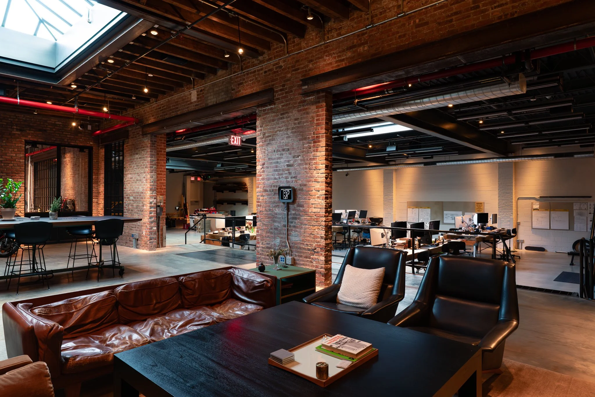 Open office space with exposed brick walls, modern furniture, multiple workstations, and large skylights letting in natural light.
