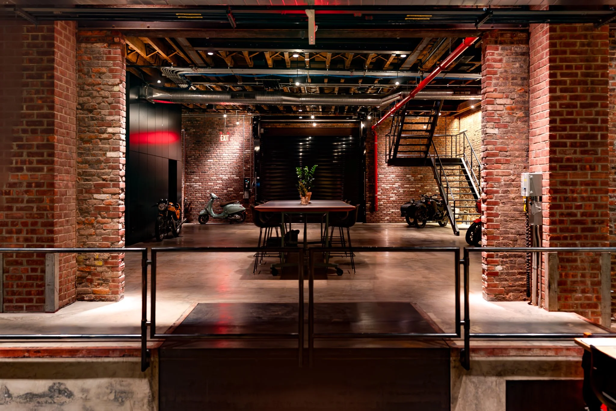 Interior view of an industrial-style building with exposed brick walls, a staircase, scooters, a table with seating, pipes on the ceiling, and an illuminated exit sign.