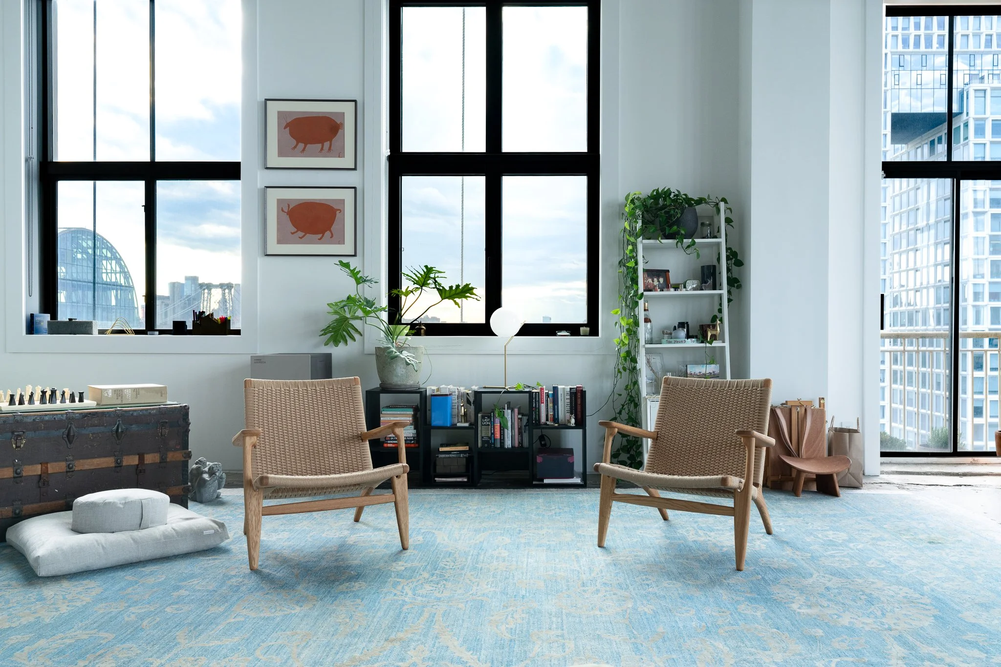 Interior of a modern high-rise apartment with large windows showing cityscape views, white walls, houseplants, a bookshelf with books and decorative items, and tan seating furniture on a blue patterned rug.