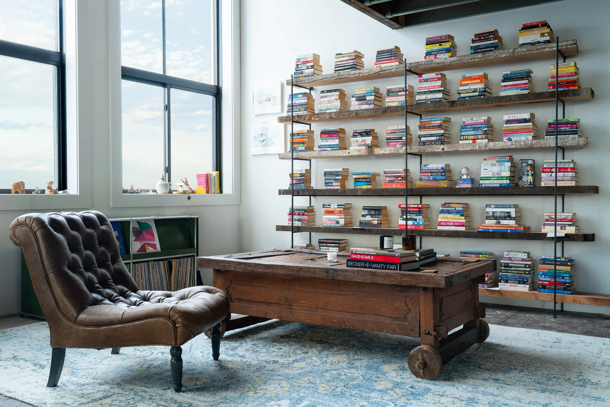 Interior of a room with large windows, a leather armchair, a wooden table, and a tall bookshelf filled with books.