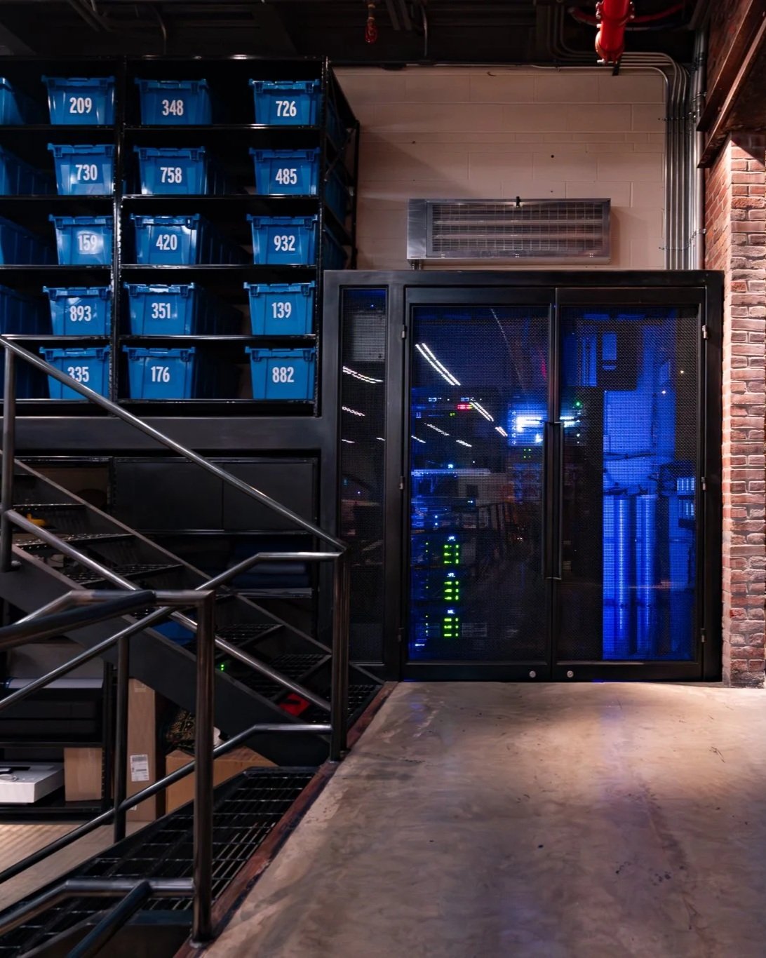 Industrial storage area with blue labeled bins on black metal shelving, a black server rack with green and blue lights, and metal stairs with railings leading up.