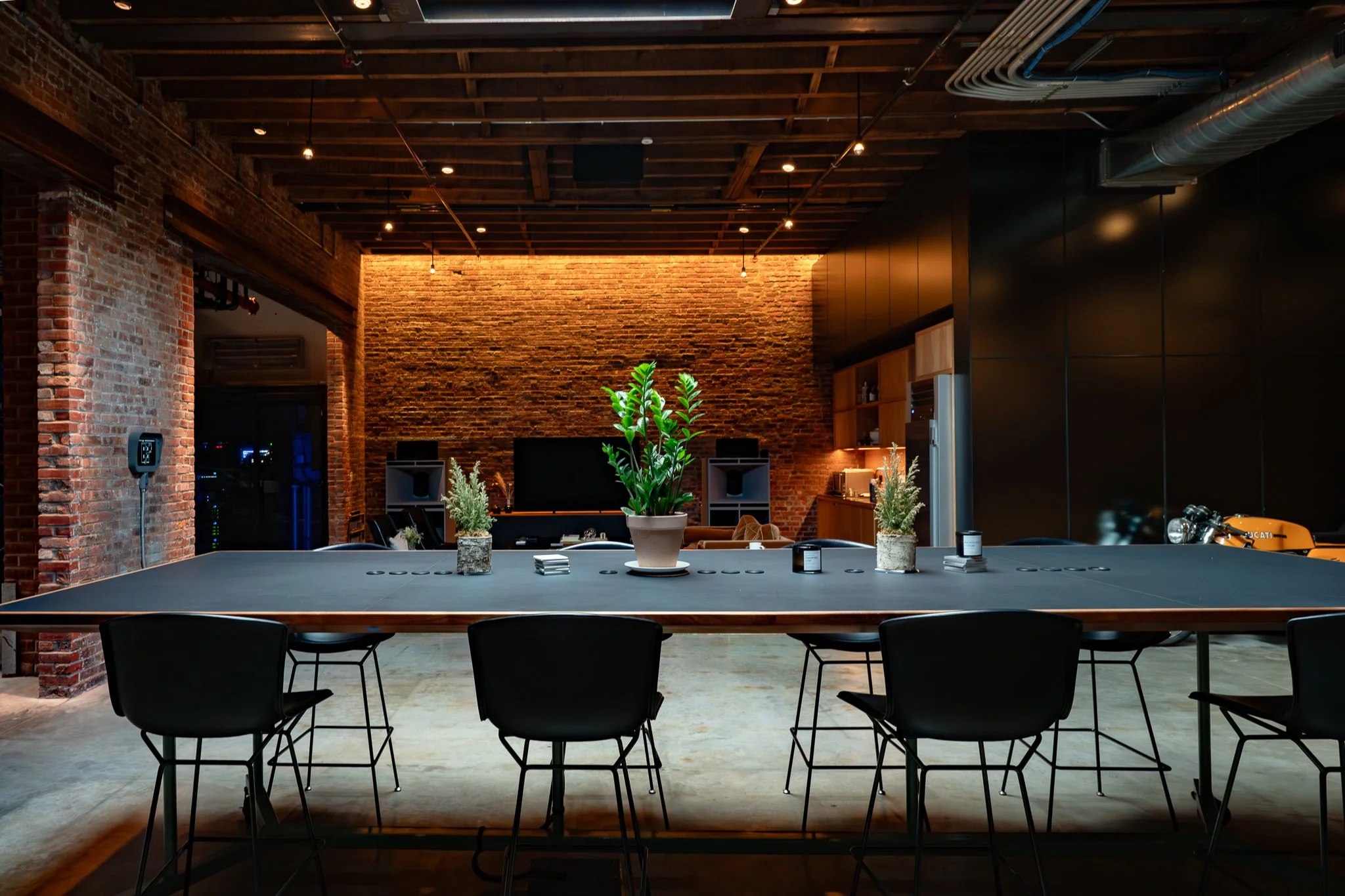 A modern industrial-style meeting room with a large rectangular table, black chairs, brick walls, potted plants, and a wooden ceiling with exposed pipes and lighting.
