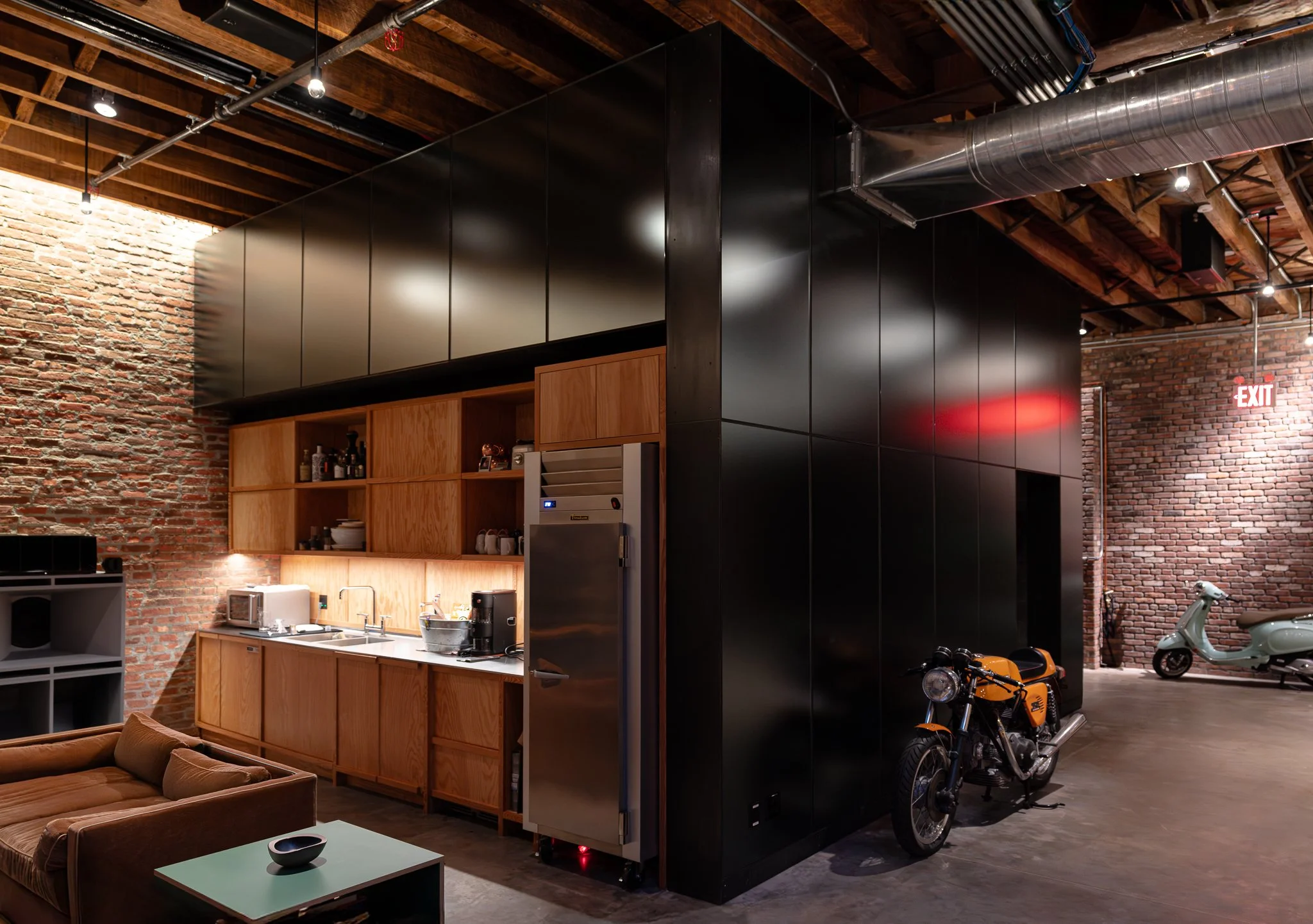 Interior view of a modern industrial-style loft with exposed brick walls and wooden ceiling beams, featuring a black partition, wooden open cabinets, a kitchen area with small appliances, and scooters parked in an open garage area.