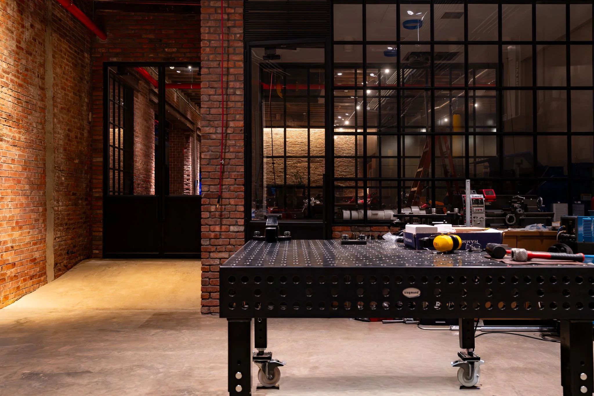 An industrial-style workspace with exposed brick walls and large glass windows, featuring a black workbench with tools and equipment.
