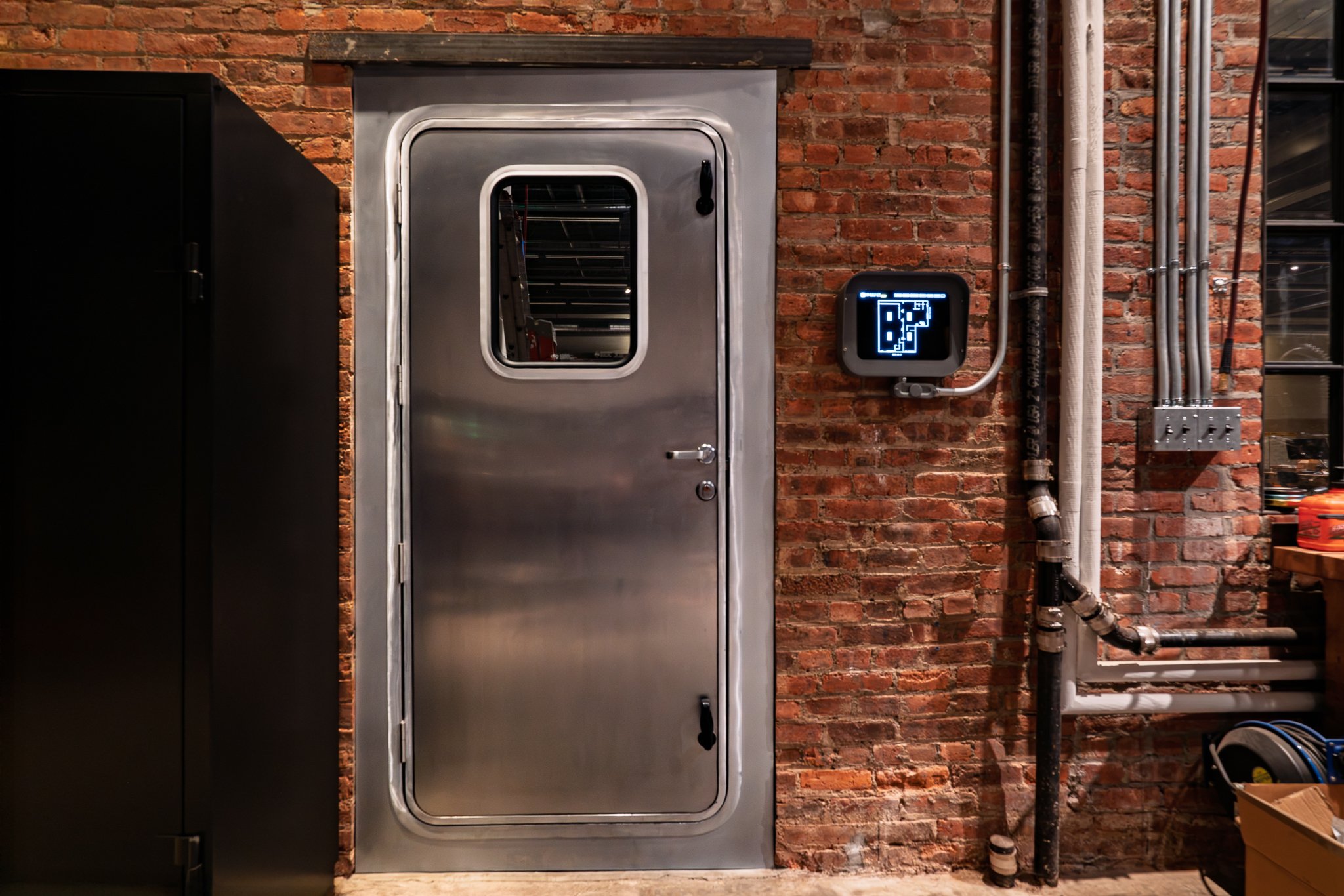 Industrial brick wall with a metal door and a digital control panel showing a diagram labeled 'B2' and temperature readings, connected to pipes and electrical wiring.