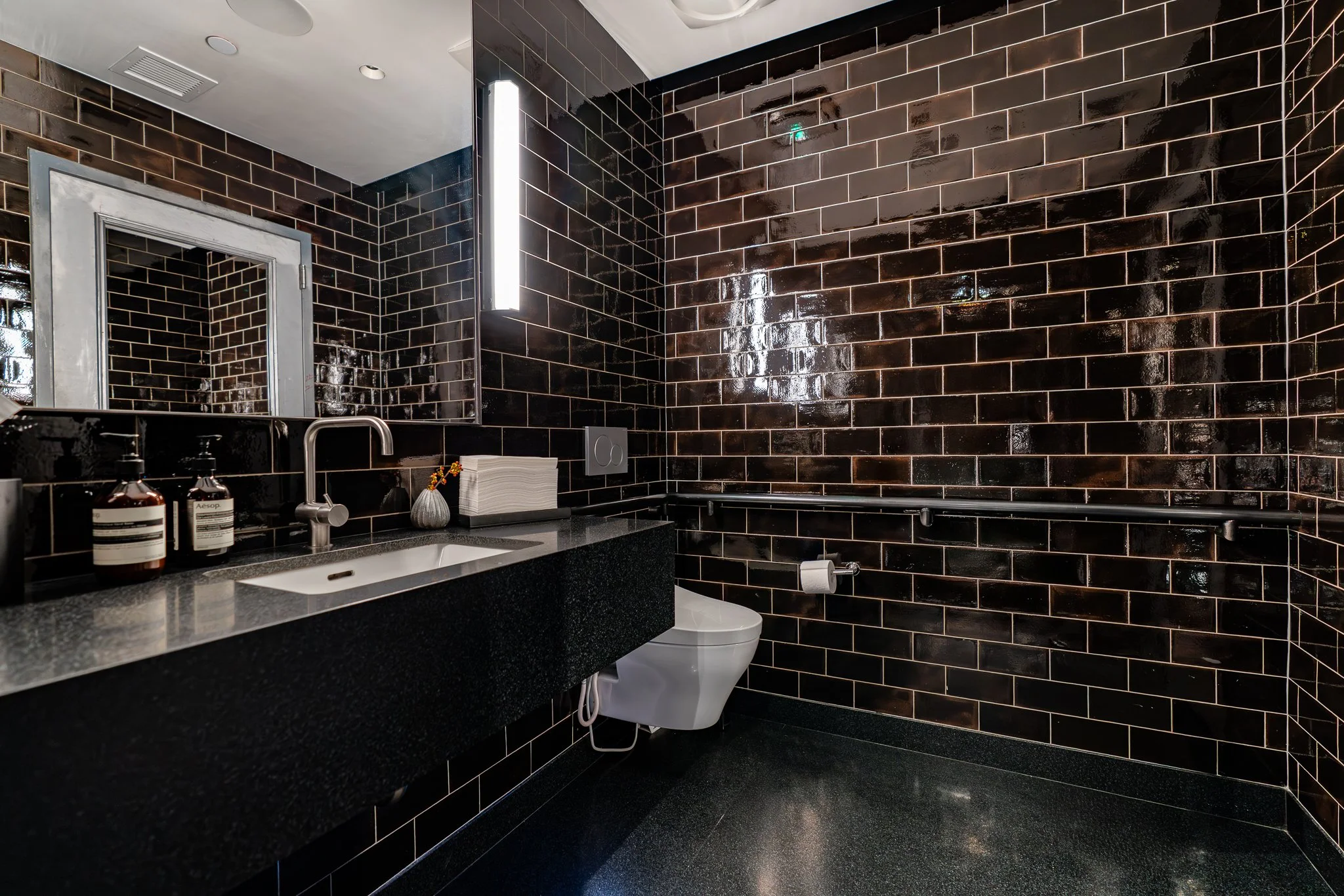 Modern bathroom with black glossy tile walls, white toilet, black vanity with a sink, various toiletries, a mirror, and sleek lighting.