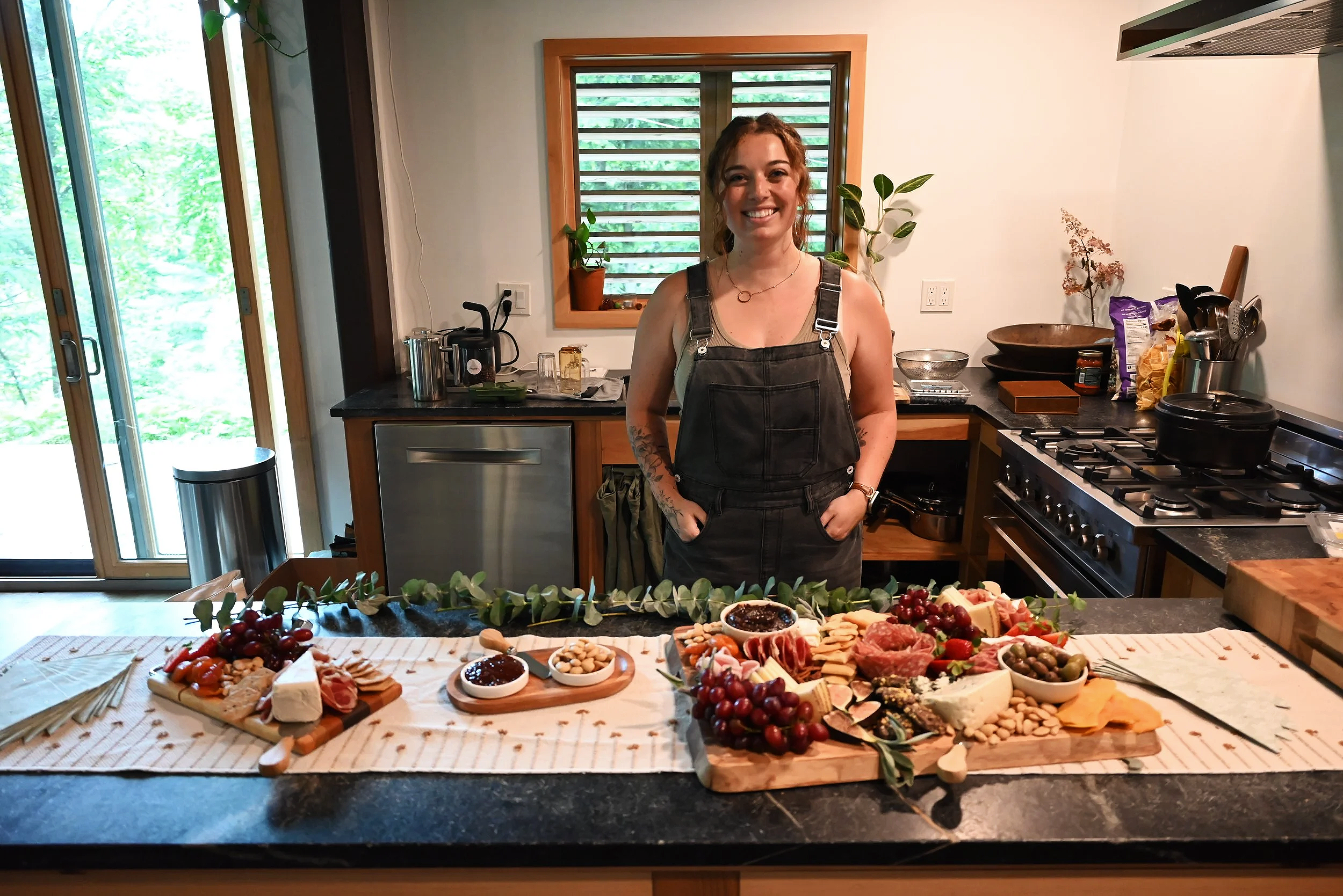A woman stands behind a food display at a kitchen counter, smiling at the camera. The counter is filled with various cheeses, grapes, and other appetizers. The kitchen has a window with wooden trim and plants. There is a stove, a small refrigerator, and kitchen utensils visible.