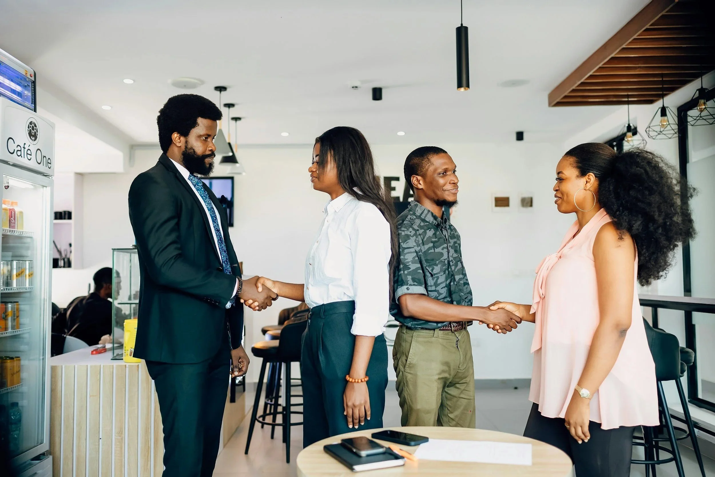 Two women and two men shake hands in a modern cafe or office space. The man on the left wears a suit, and the woman he is shaking hands with wears a white blouse. The other woman, with curly hair, wears a pink sleeveless blouse. The man next to her wears a camouflage shirt. Other customers are seated in the background.