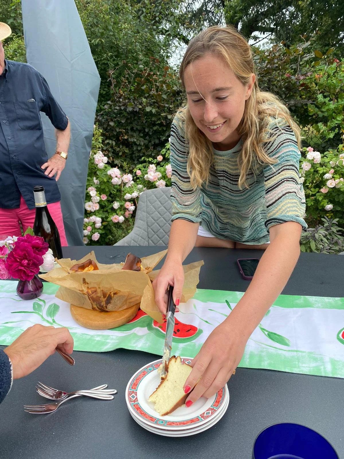 A young woman is serving herself a slice of cake at a table decorated with a watermelon-themed tablecloth, flowers, and food, outdoors in a garden with blooming pink roses.