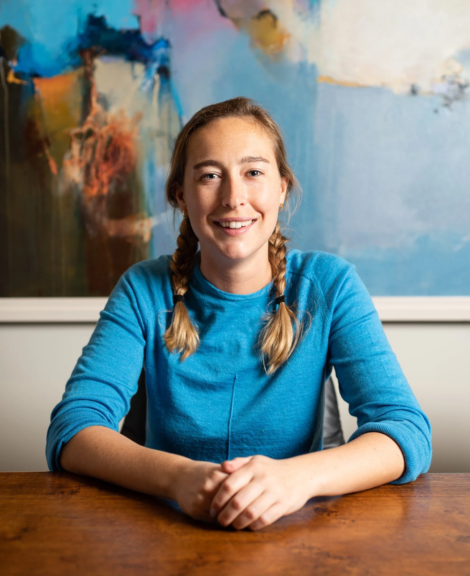 A young woman with braided hair smiling, sitting at a wooden table with her hands folded in front of her, in front of an abstract blue and brown painting.