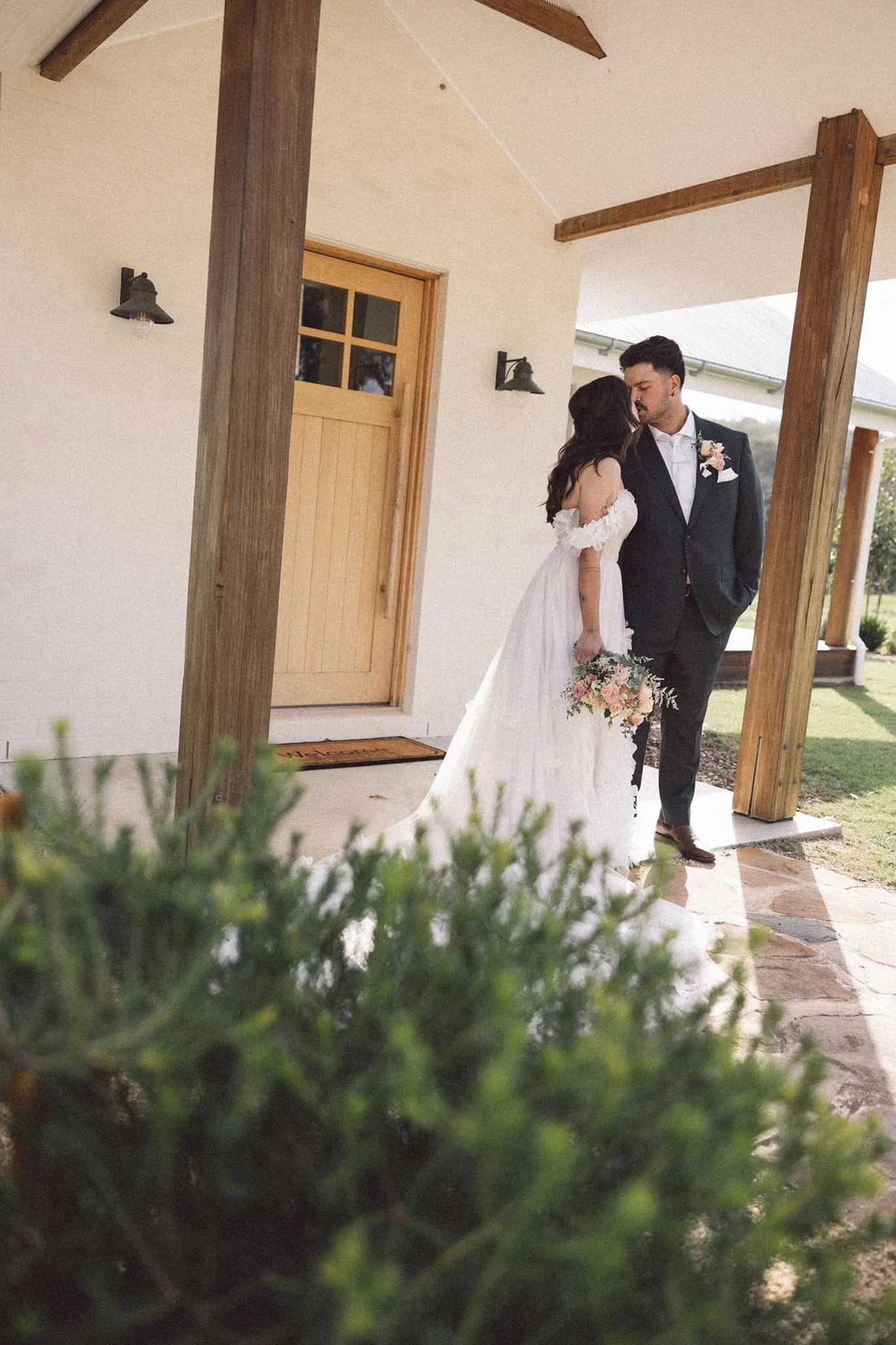 A bride and groom share a kiss outside a house with a wooden door, seen through some green plants in the foreground.