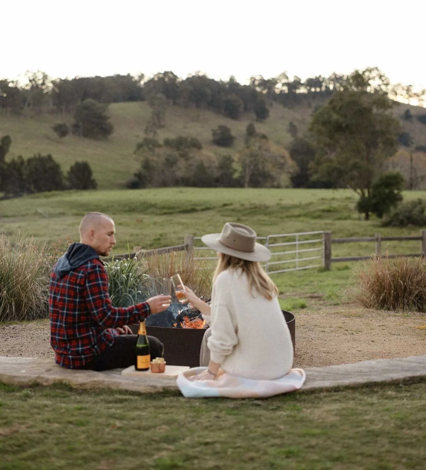 A man and woman having a picnic outdoors near a fire pit with drinks, in a grassy area with fences and rolling hills and trees in the background.