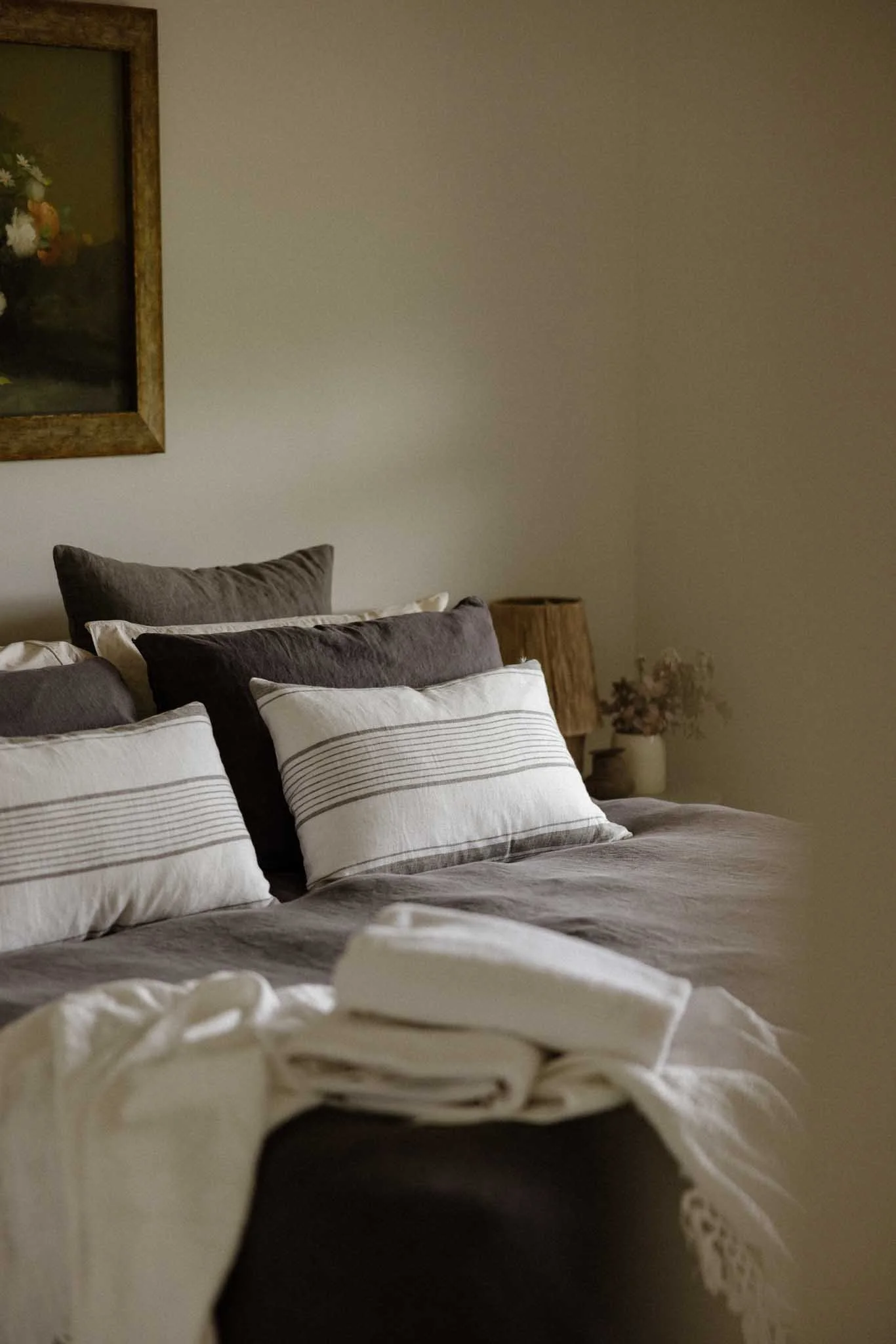 A neatly made bed with white and brown striped bedding and multiple pillows, including white pillows with thin black stripes and darker pillows, in a cozy bedroom with a small wooden side table and a framed floral painting on the wall.