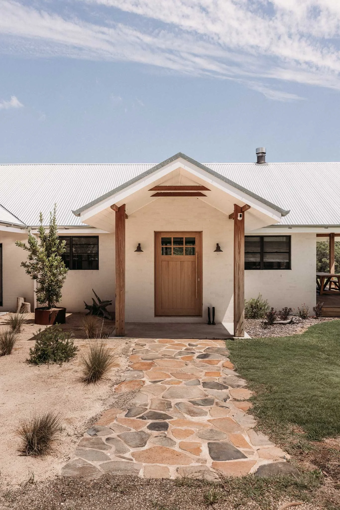 Front view of a modern house with a gable roof, wooden columns, and a stone pathway leading to the front door, surrounded by desert landscaping.