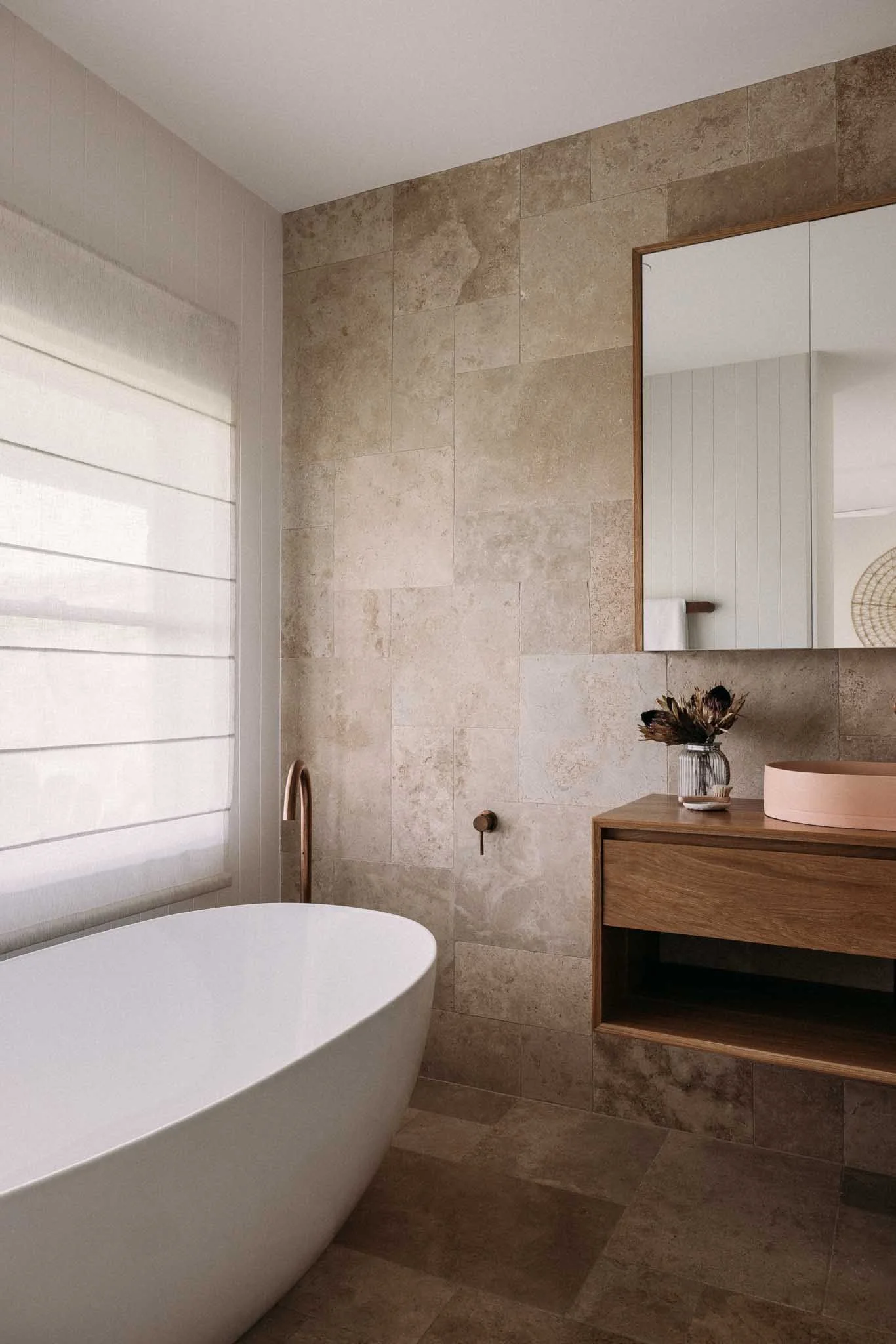 A bathroom with a freestanding bathtub, beige tiled walls, a large mirror with a wooden frame, a wooden cabinet with a vase of dried flowers on top, and a white window blind.