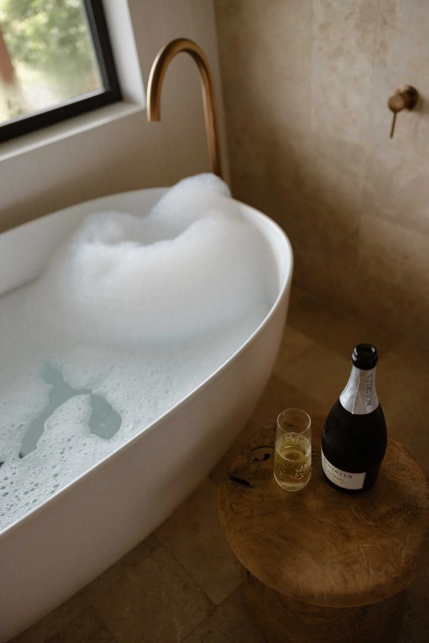 A bathtub filled with bubbles, a window showing greenery outside, a corked bottle and a glass of champagne on a wooden table next to the bathtub.
