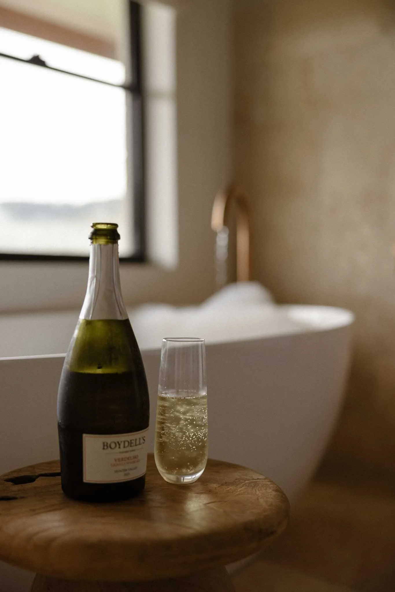 A bottle of Boydell's sparkling wine and a filled champagne flute on a round wooden table, with a blurred window and wall in the background.