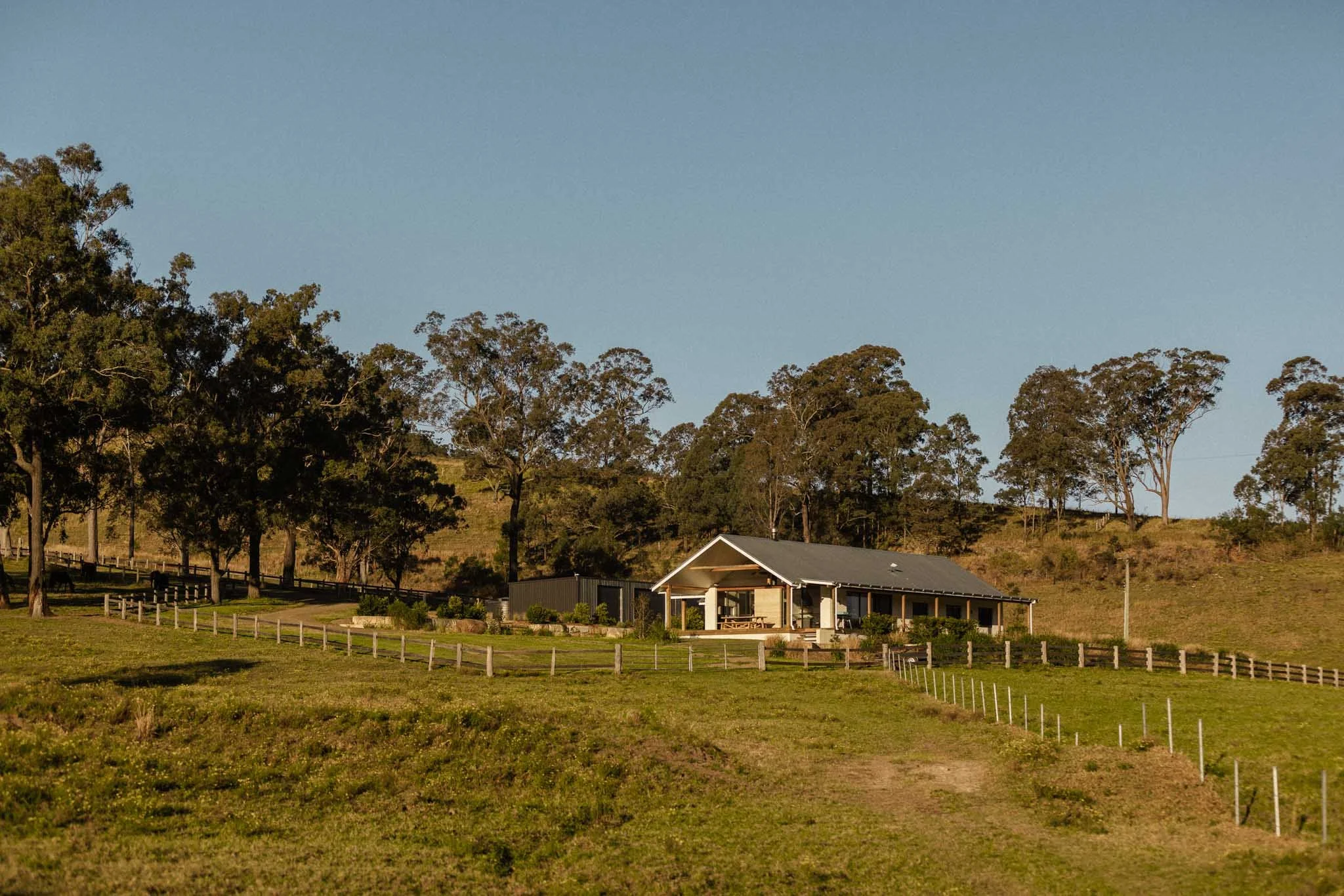 Single-story house with a gray roof and covered porch, surrounded by green landscape with trees and fencing, under a clear blue sky.