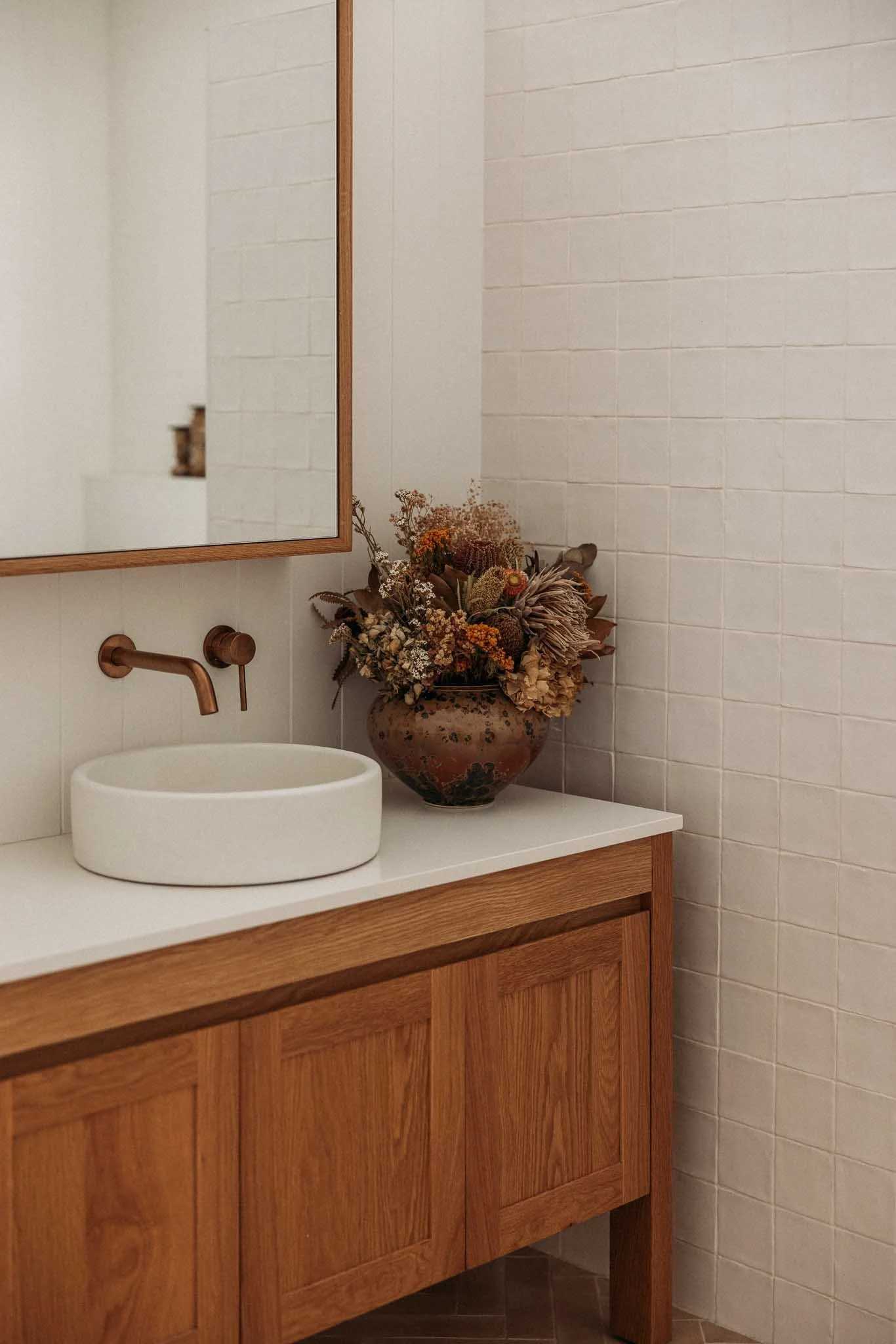 A bathroom with a wooden vanity, white countertop, round white vessel sink, bronze faucet, a large mirror with wooden frame, a bouquet of dried flowers in a pot, and a beige tiled wall.