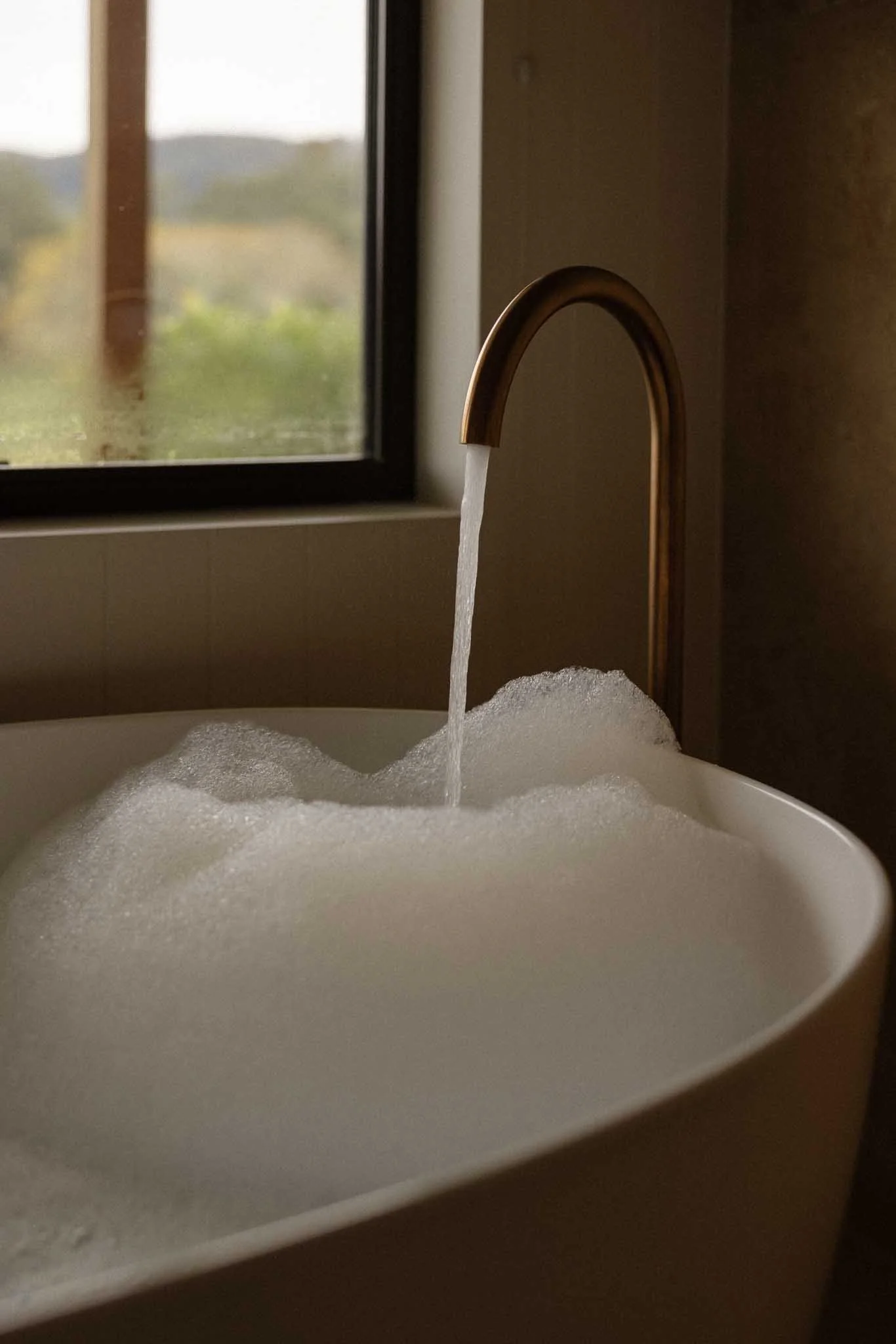 A close-up of a metallic gold faucet with water running into a white sink filled with soap bubbles, near a window with natural light.
