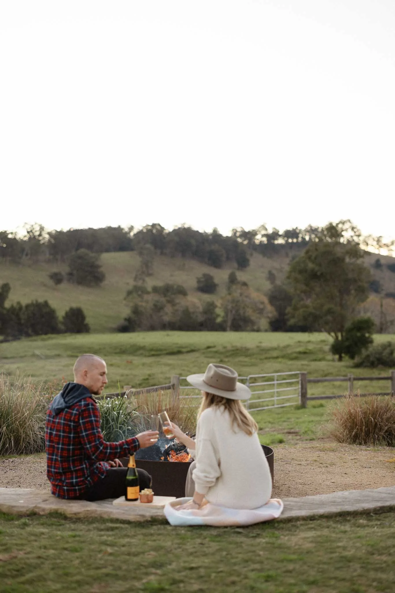 Two people sitting on a blanket outdoors near a fire pit, toasting with drinks, with a rustic fence and green rolling hills in the background.