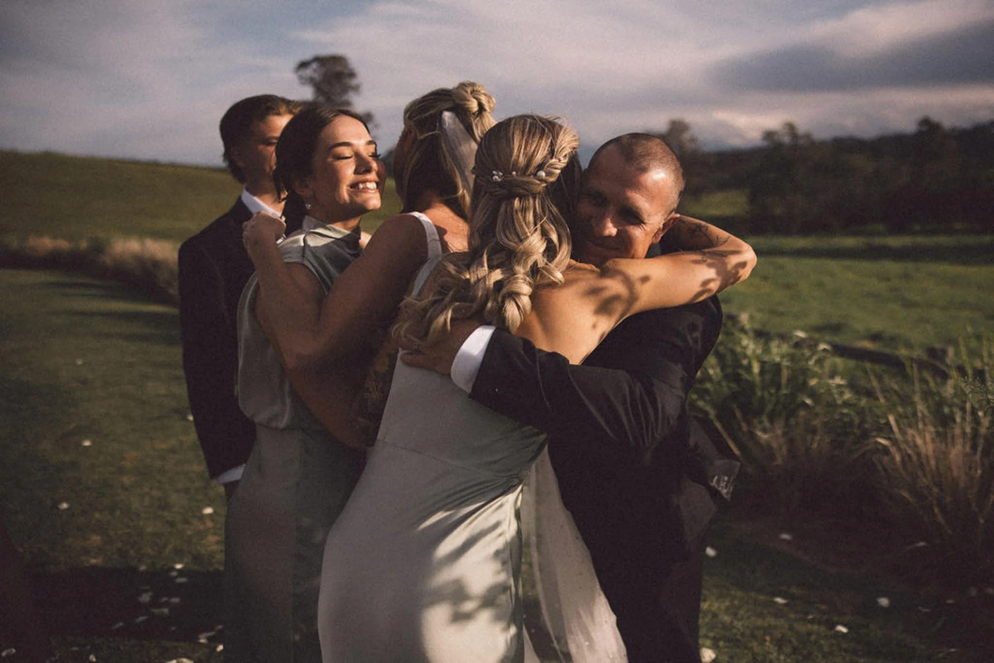 People hugging outdoors during sunset, with a grassy landscape and trees in the background.