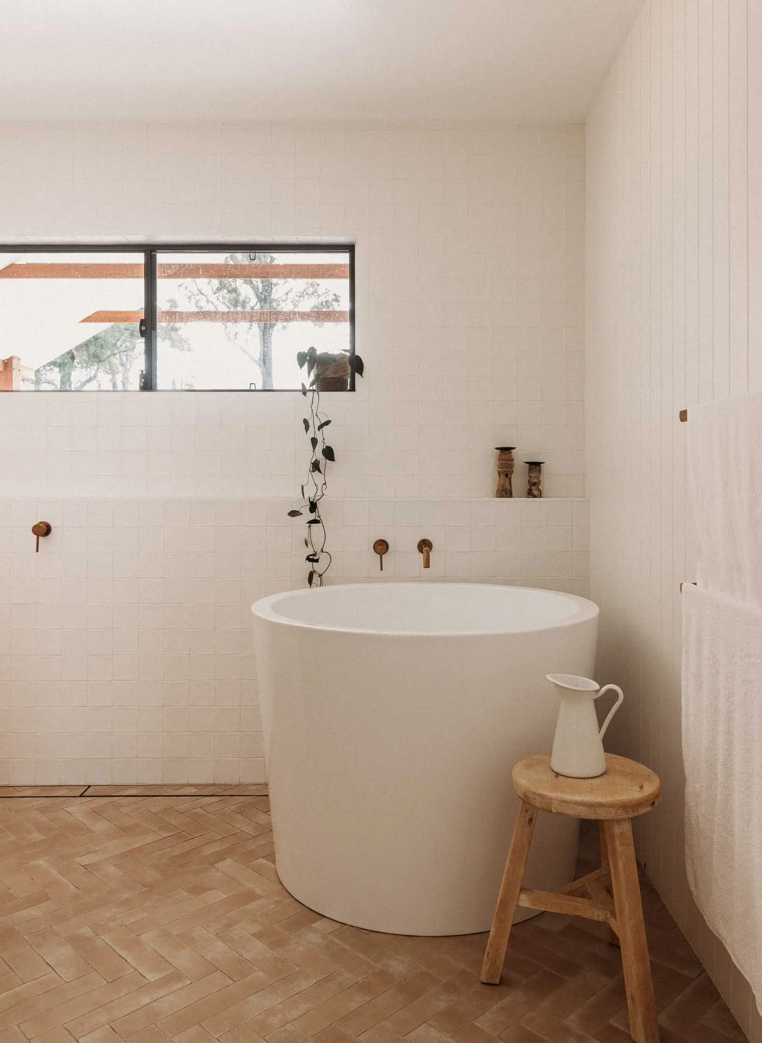 Minimalist bathroom with a tall white freestanding bathtub, a small wooden stool with a white pitcher vase, beige tiled floor, white tiled wall, window with outdoor view, and decorative walking sticks on wall.