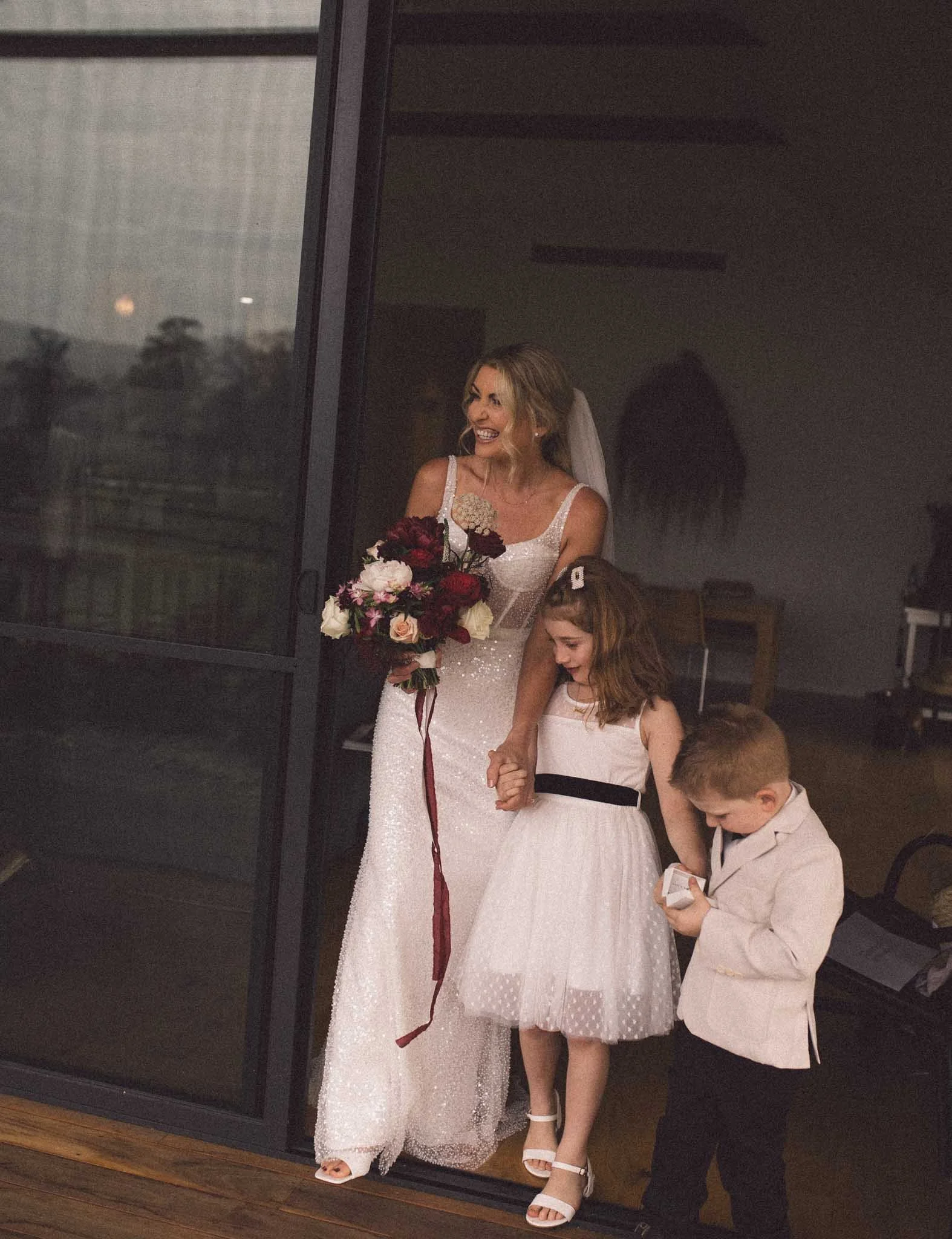 A bride in a white, sparkly wedding dress holding a bouquet, smiling behind a glass door, with three children dressed in light-colored formal attire, standing outside and looking down.