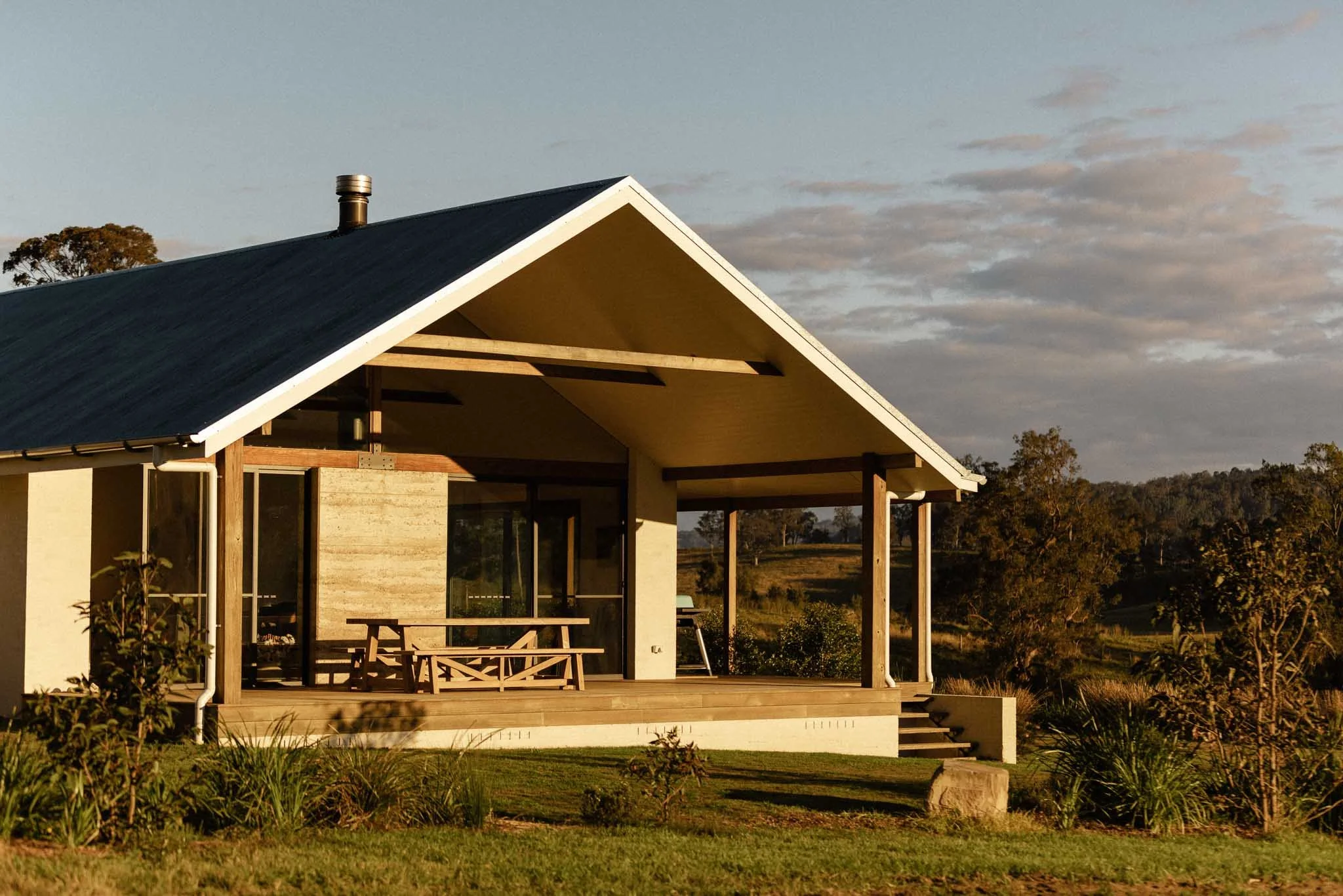 A modern house with a sloped roof and a patio, surrounded by grass and trees, during sunset.