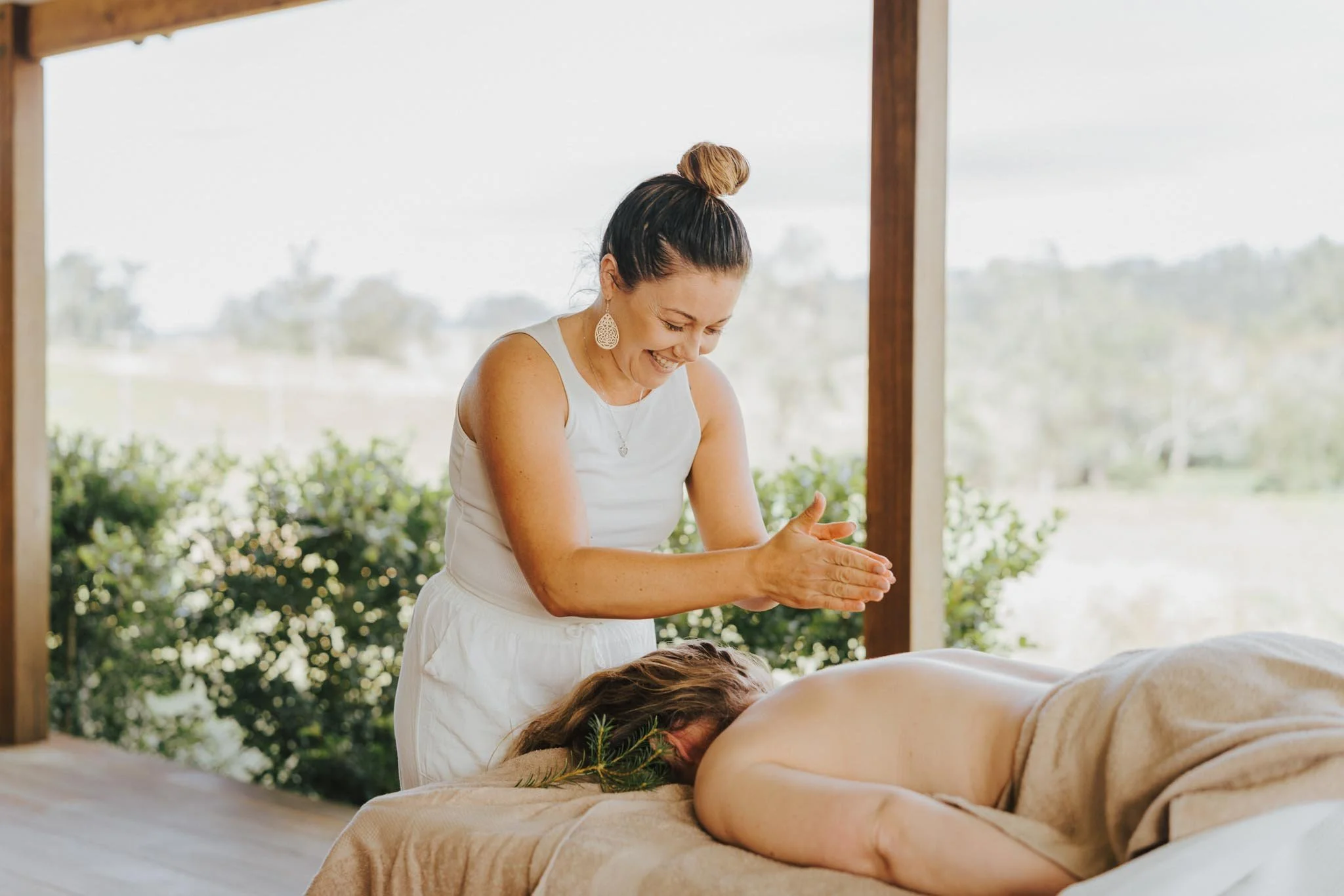 A woman in a white dress giving a massage outdoors under a wooden structure, with greenery and trees in the background.
