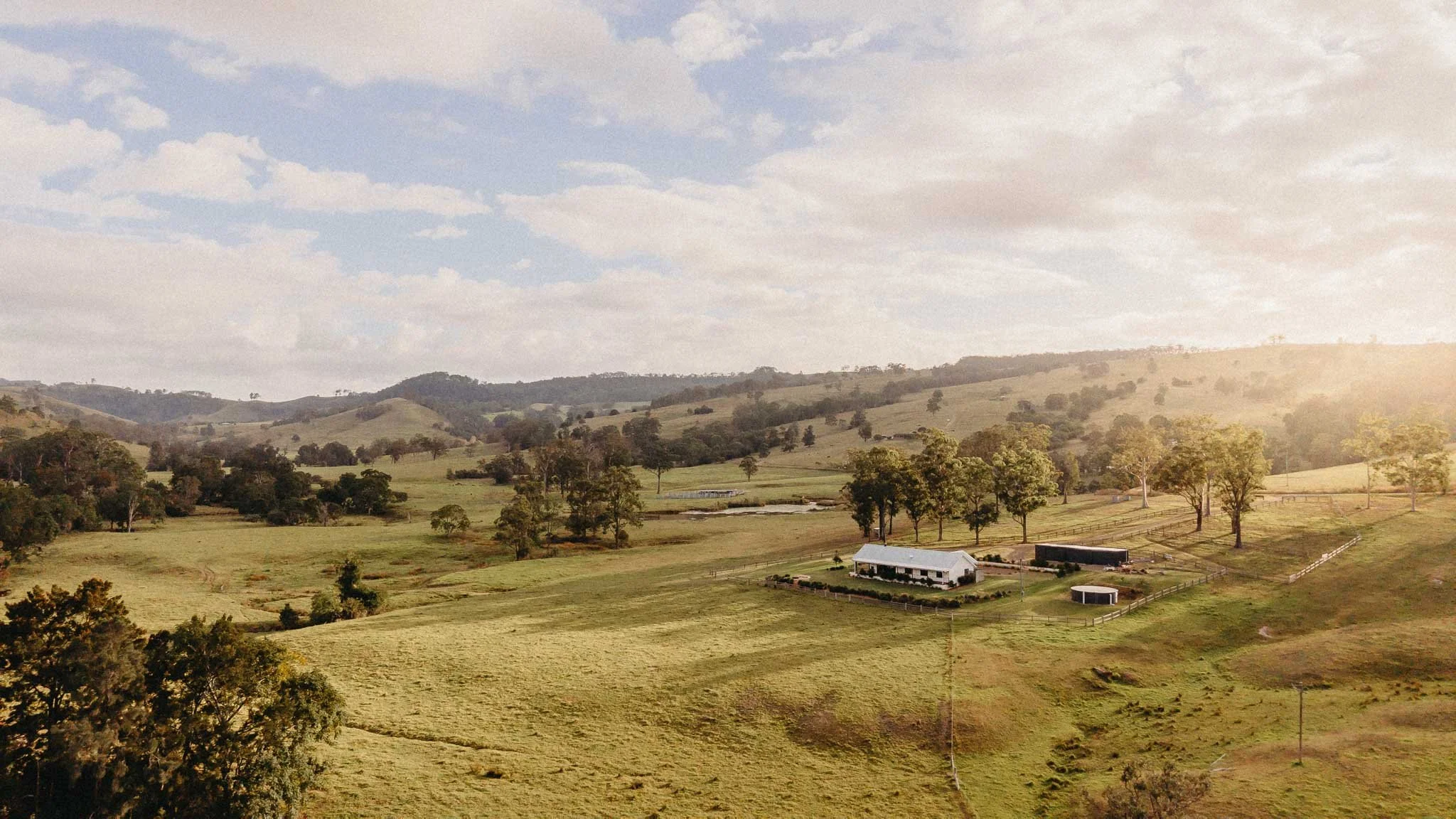 Scenic view of rolling green hills, scattered trees, a farmhouse, a pond, and a fenced yard under a partly cloudy sky with sunlight.