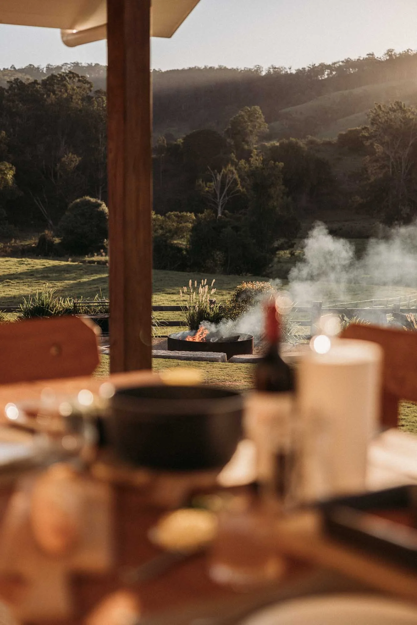 A scene from an outdoor patio with a fire pit emitting smoke, surrounded by greenery and trees, with hills in the background, viewed through a partially obstructed view with out-of-focus objects in the foreground.