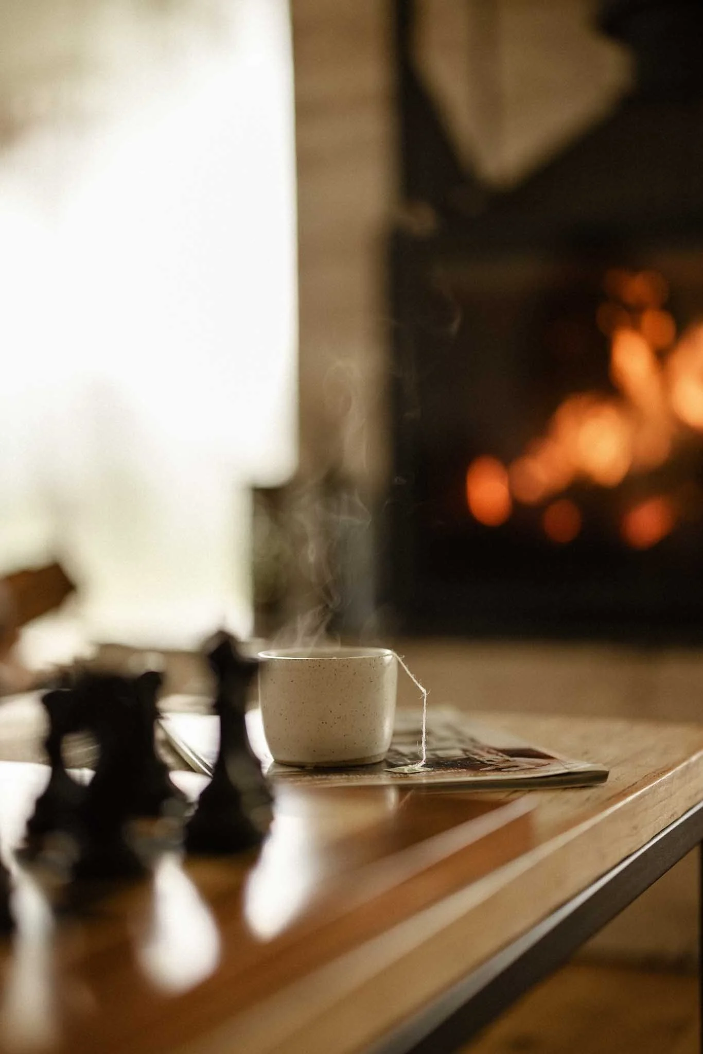 Close-up of a steaming cup on a wooden table with a blurred chessboard in the foreground and a fireplace with glowing fire in the background.