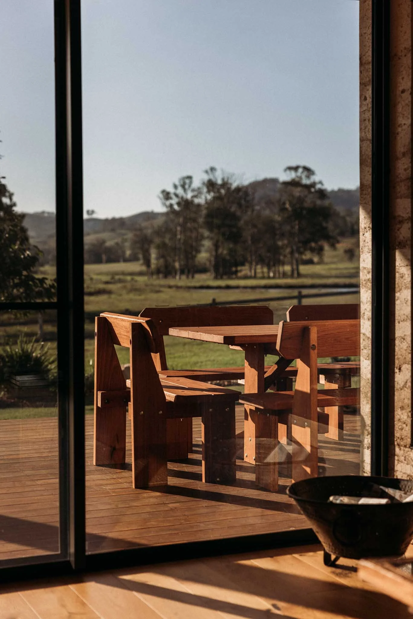 A view of a wooden outdoor patio with chairs and a table, seen through a sliding glass door. The landscape in the distance includes trees and rolling hills under a clear sky.
