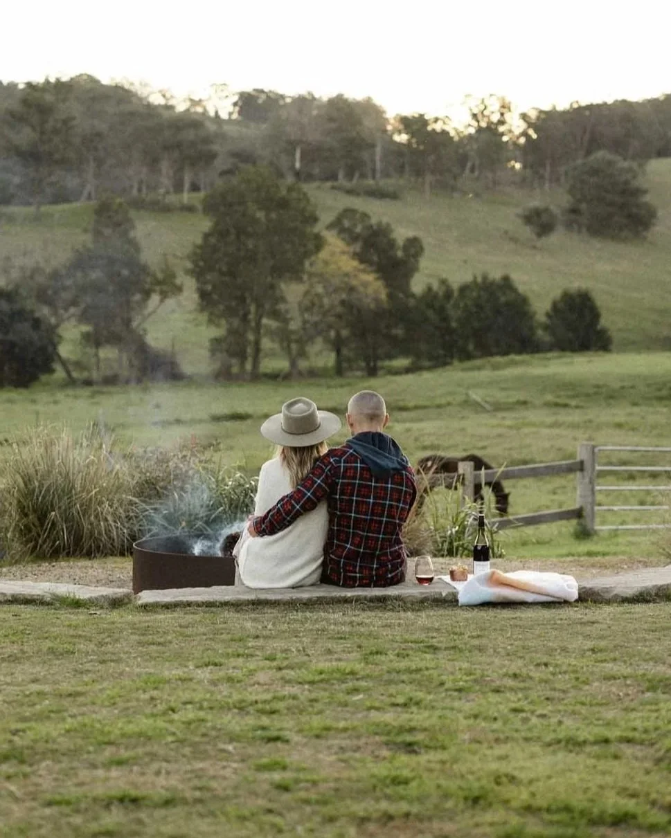 Couple with arms around each other sitting by a campfire outdoors, enjoying a scenic countryside with green hills, trees, and grazing horses, in the early evening.