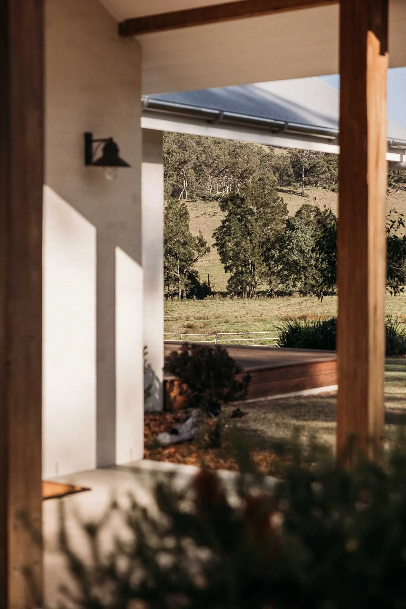 Photo taken from a porch or patio, looking out toward a rural landscape with trees and open grassy fields, with part of the house visible in the foreground.