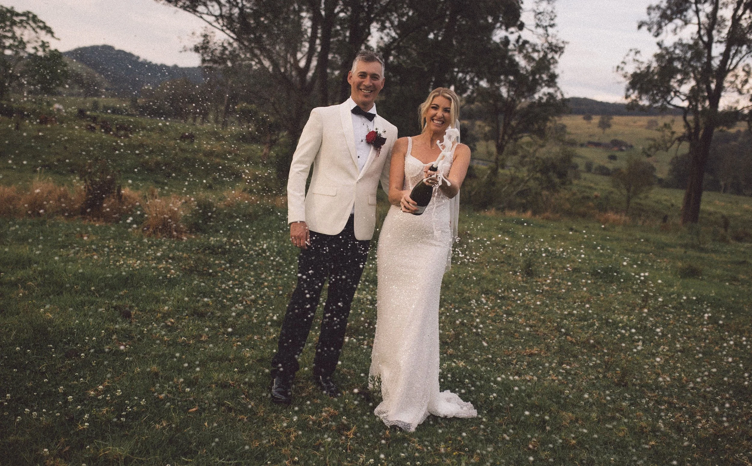 A bride and groom are kissing in a field with horses nearby, under a cloudy sky, during what appears to be a wedding photo shoot.