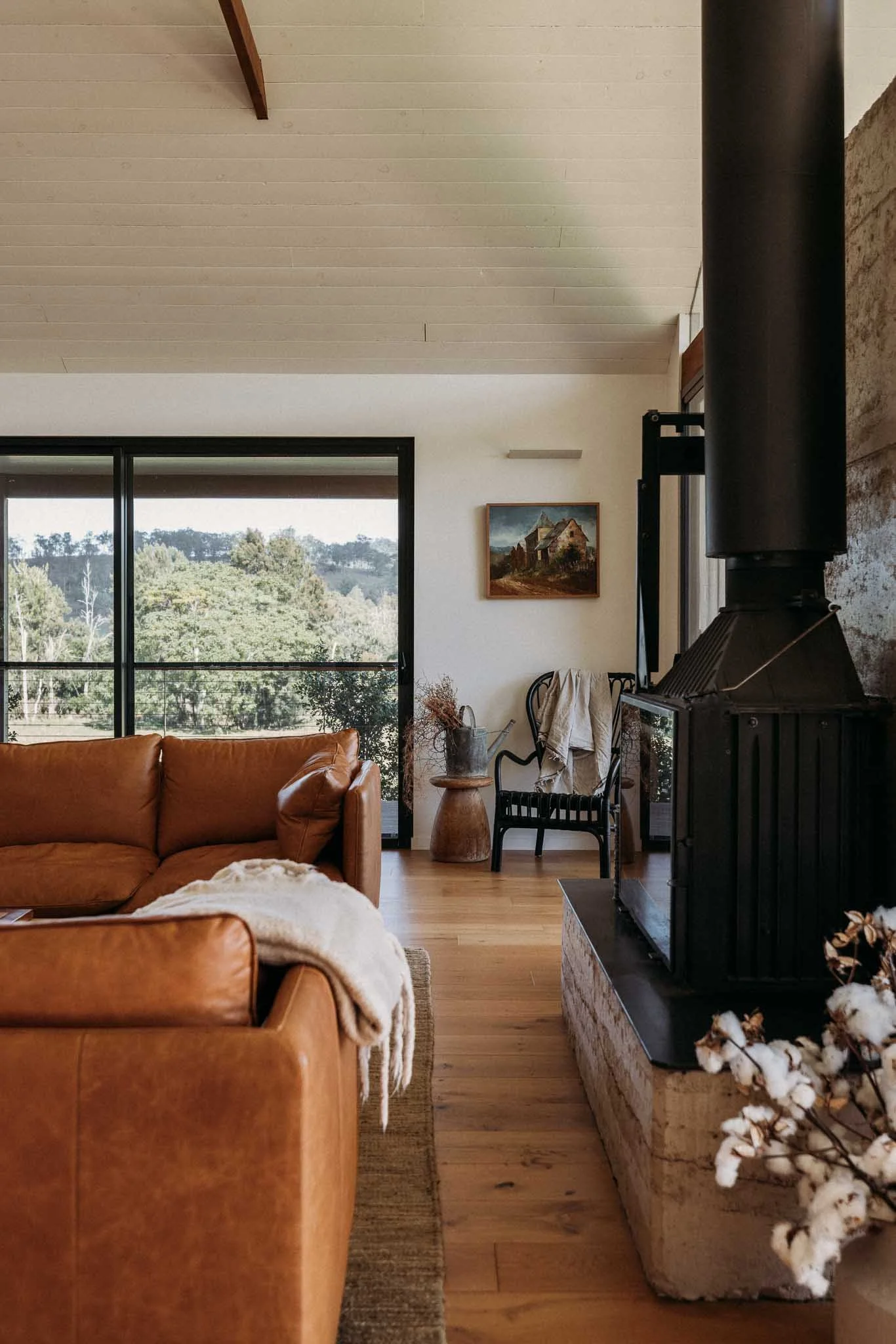 A living room with leather sofas, a window showing a view of trees, a painting on the wall, and a fireplace with cotton branches.