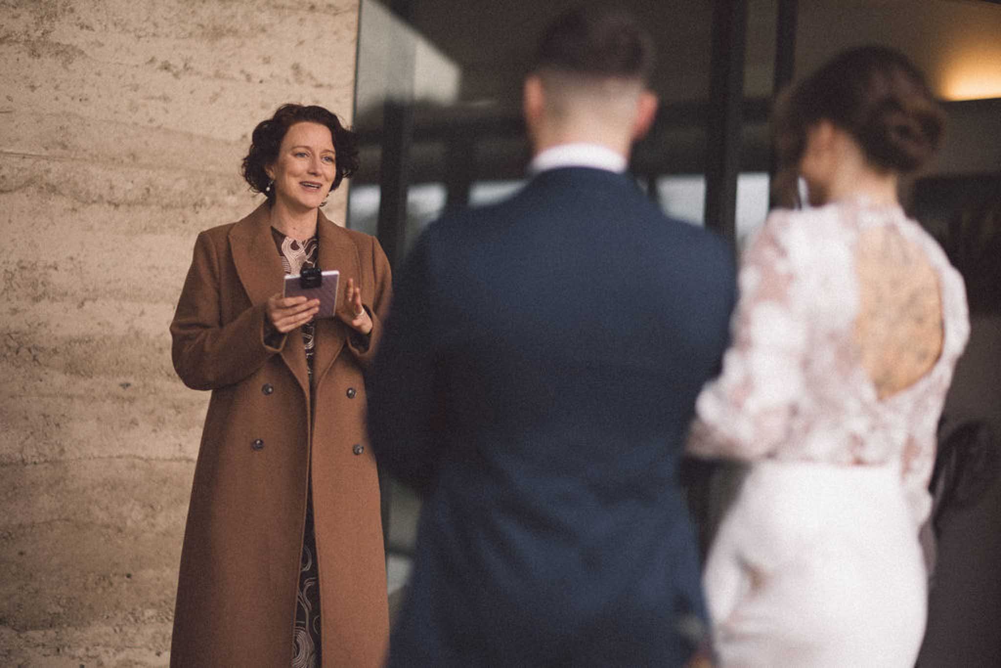 A woman with curly dark hair wearing a brown coat talking to two people, a man and a woman, in an indoor setting with a stone wall and a dark background.