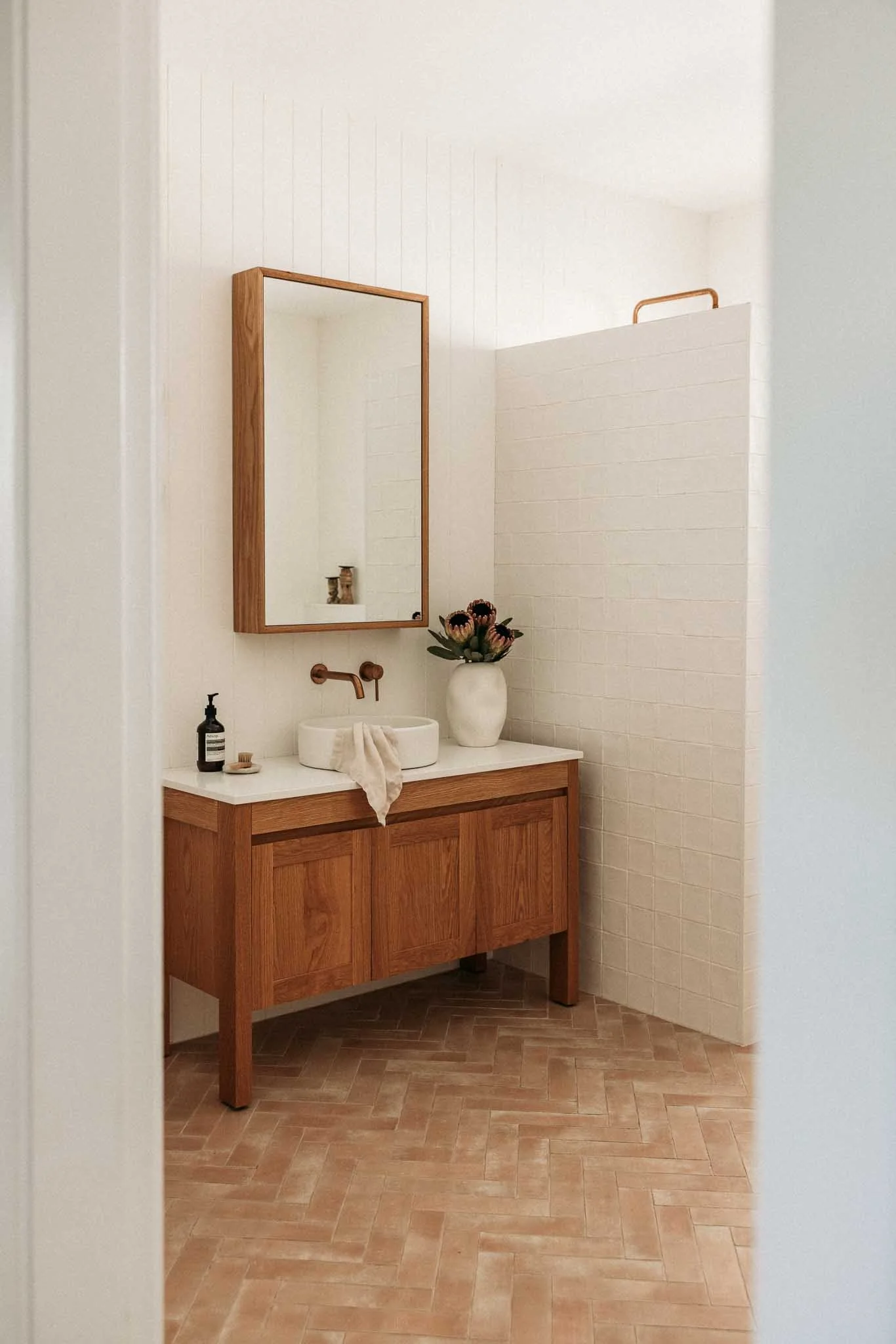 Bathroom vanity with a vessel sink, a mirror with a wooden frame, a bottle of soap, a small plate, a towel, a vase with flowers, and a privacy partition.