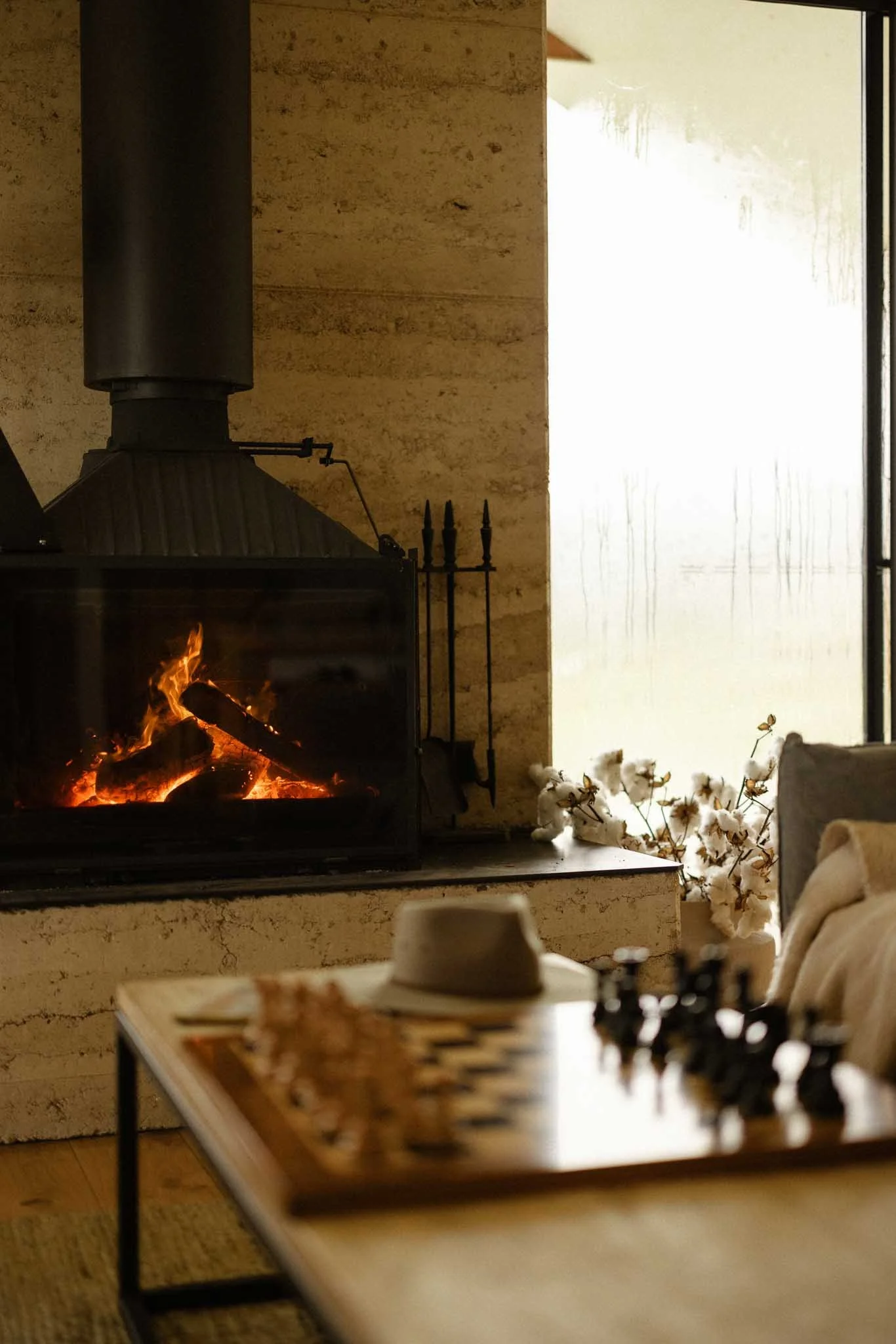 A lit fireplace in a cozy living room with cotton branches and a blurred chessboard on a table in the foreground.